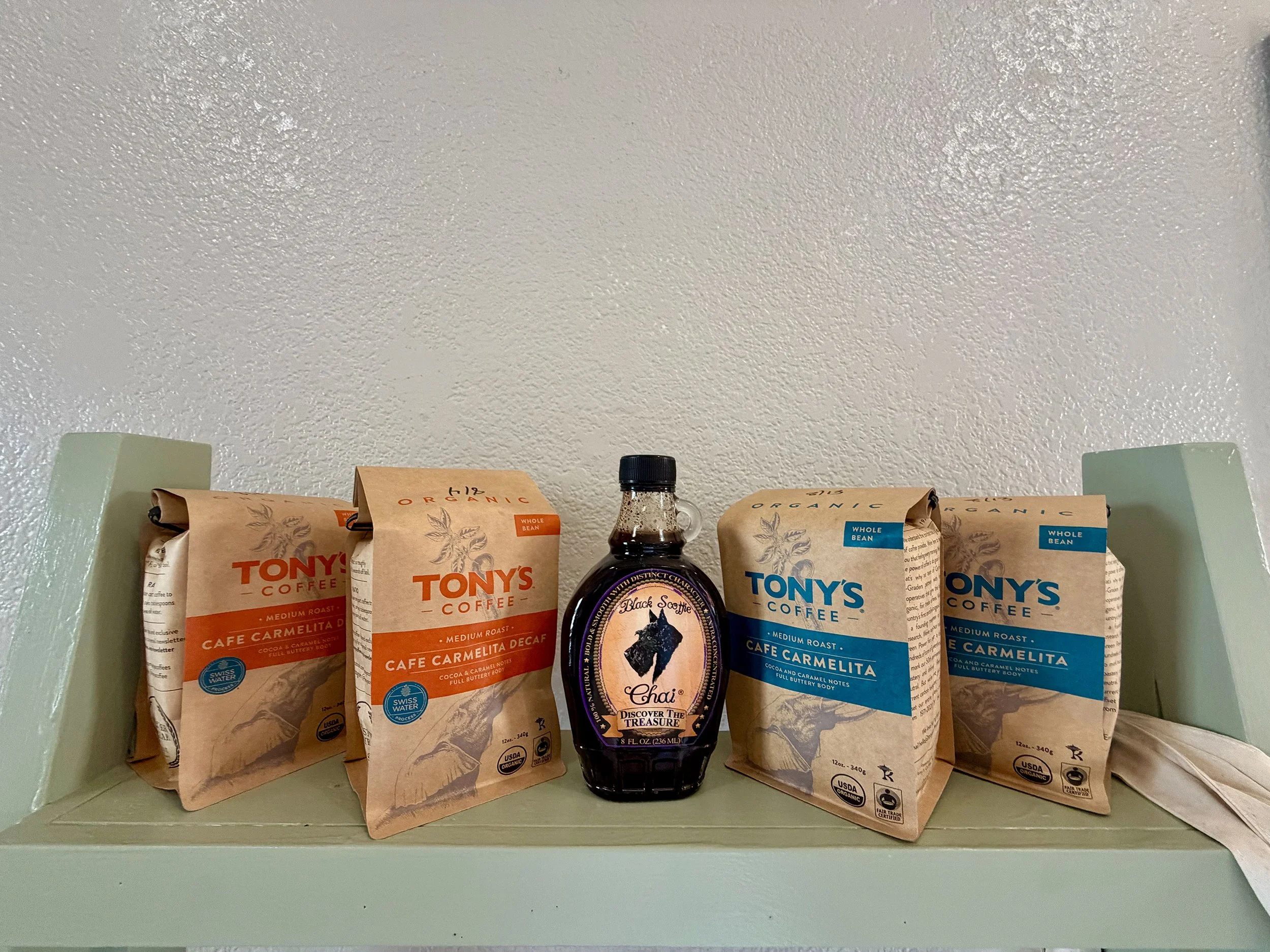 Shelf with various bags of Tony's Coffee, including Cafe Carmelita and Organic Whole Bean varieties, and a bottle of Chai syrup in the center.