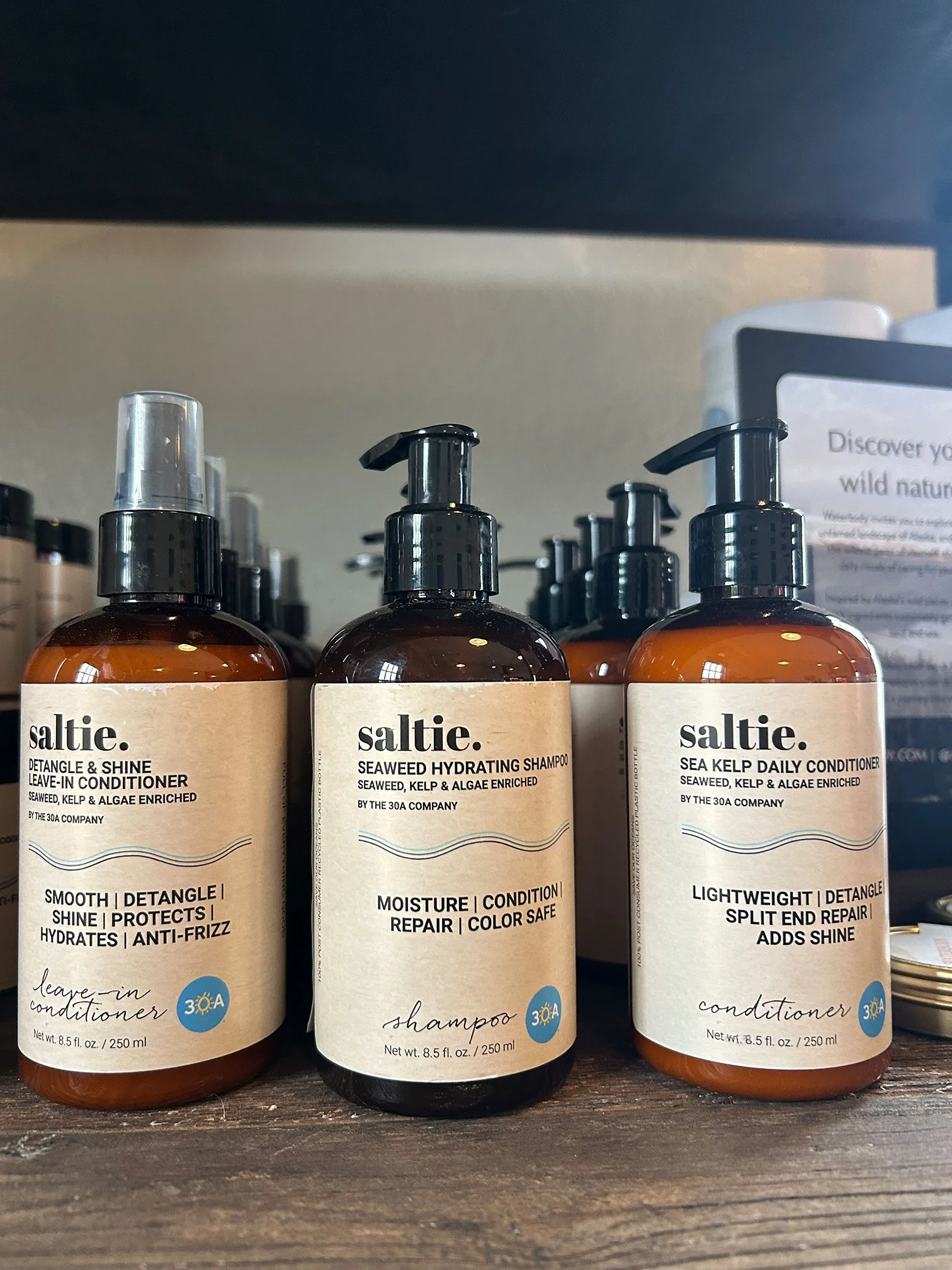 Three bottles of hair care products from the brand Saltie, including leave-in conditioner, shampoo, and conditioner, arranged on a wooden surface. The bottles are amber-colored with black pumps and beige labels.