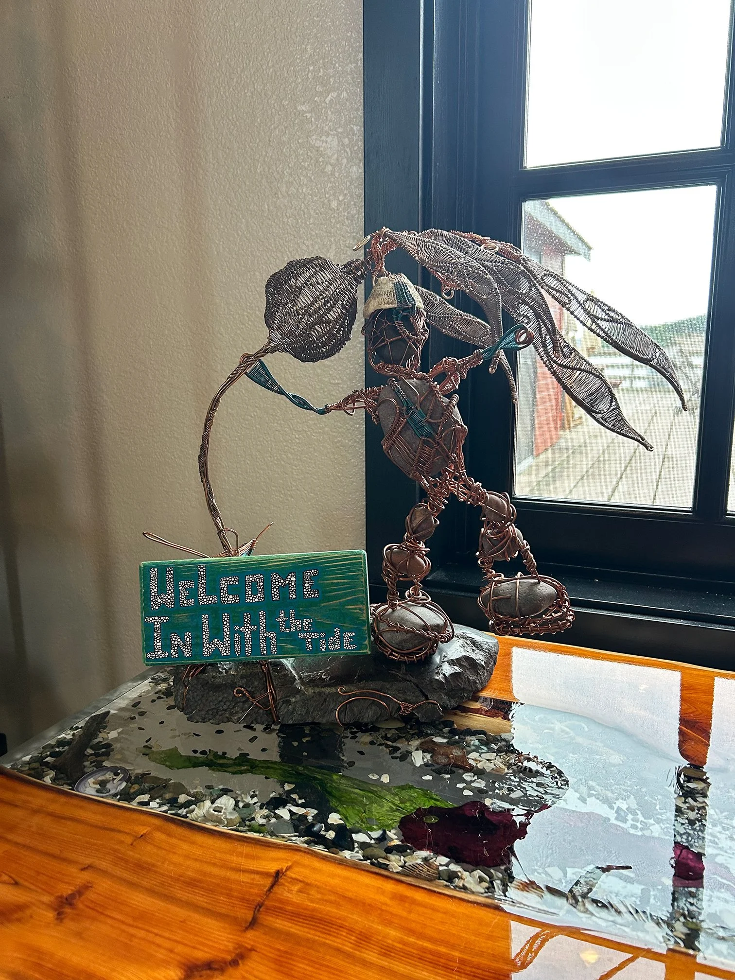 A decorative wire art sculpture of a fairy standing on a stone base near a window, accompanied by a colorful sign that says "WELCOME TO THE TIDE." The sculpture features detailed wings, hair, and clothing made of wire, with objects like shells and st