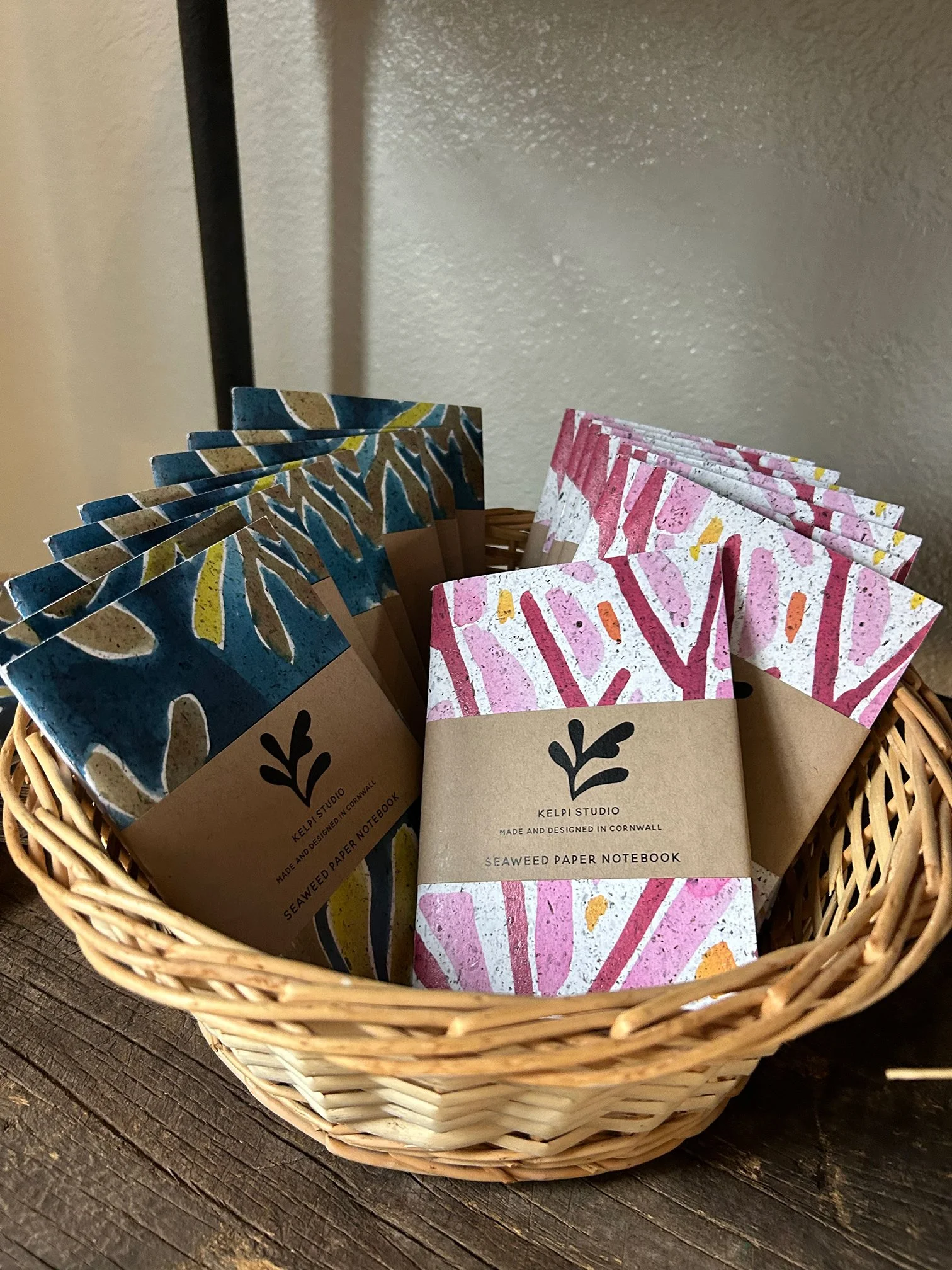 A wicker basket containing small notebooks with colorful, abstract, paper covers. The notebooks are labeled 'Seaweed Paper Notebook' from Kelpi Studio, designed and made in Cornwall, with a stylized leaf logo on the cover.