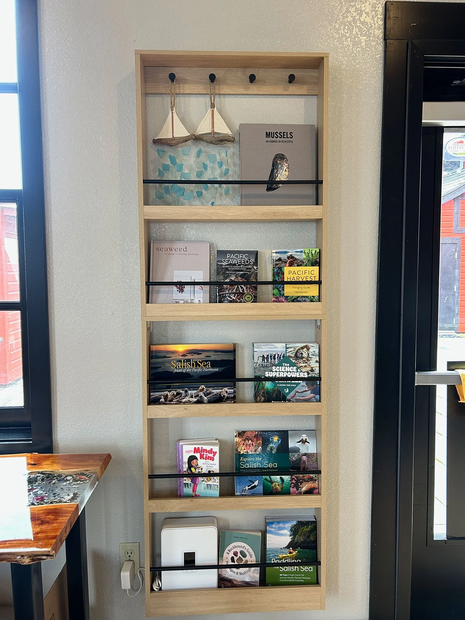 A wooden bookshelf with five shelves displays various books about nature, the sea, and kayaking, with decorative items like a small sailboat, sea shells, and decorative rocks, positioned against a wall near a window and black framing.