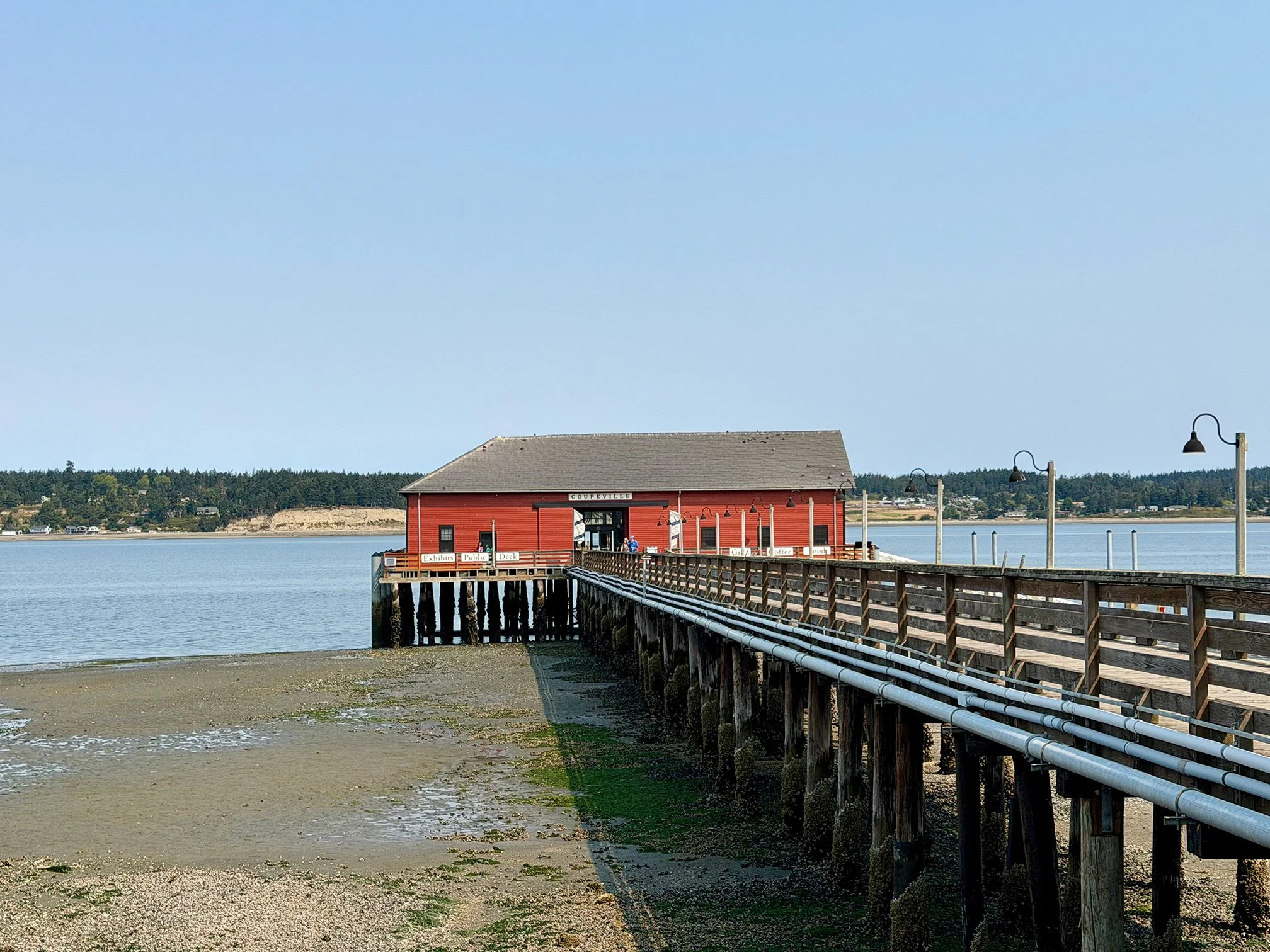 A red building on stilts extending over water with a dock and walkways, surrounded by a waterfront with some trees in the background and clear sky above.