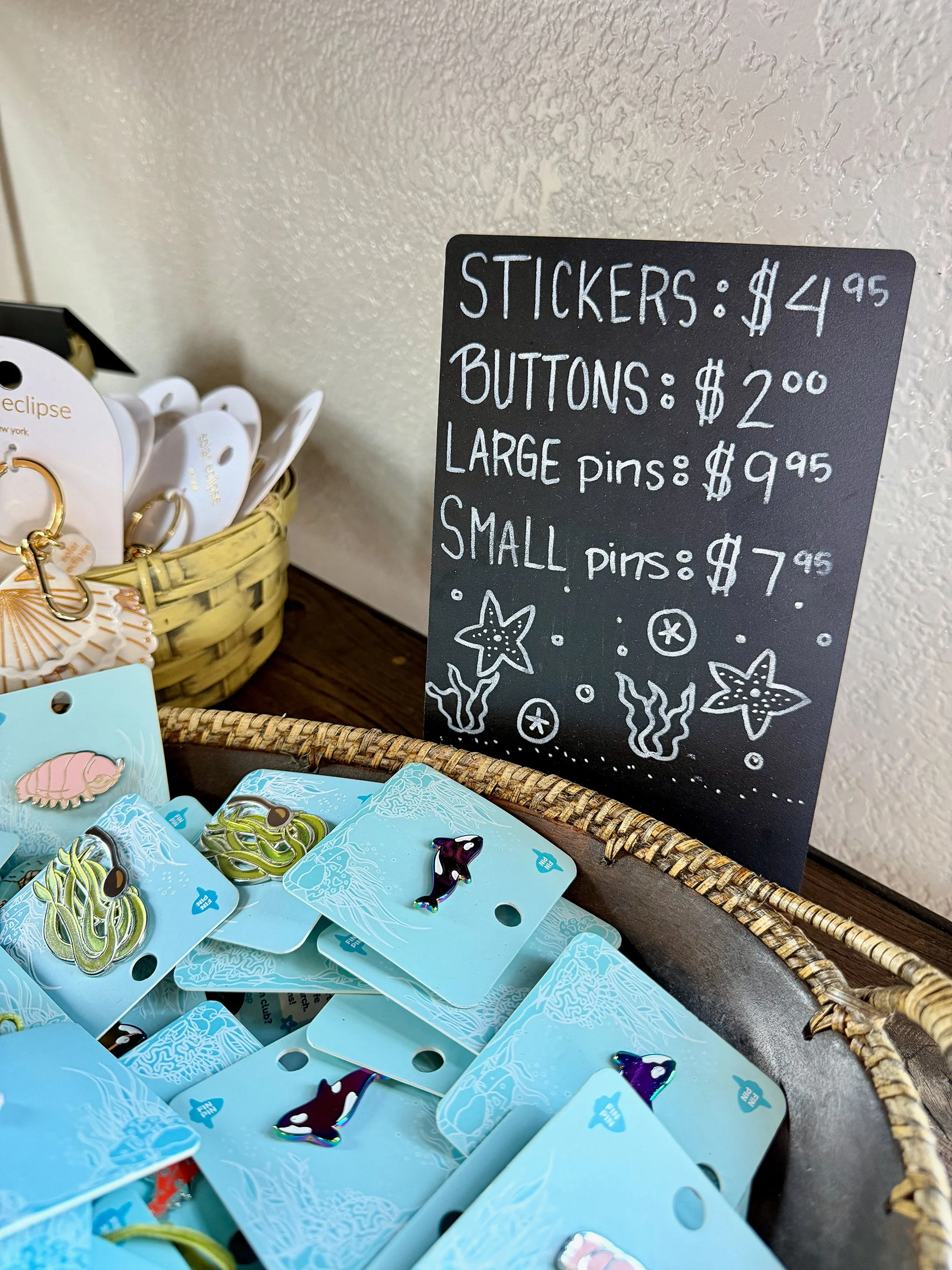 A display of small collectible pins, including a pink sea urchin and a purple dolphin, in a woven basket. A blackboard sign lists prices for stickers, buttons, and pins, with illustrations of starfish, seaweed, and bubbles.