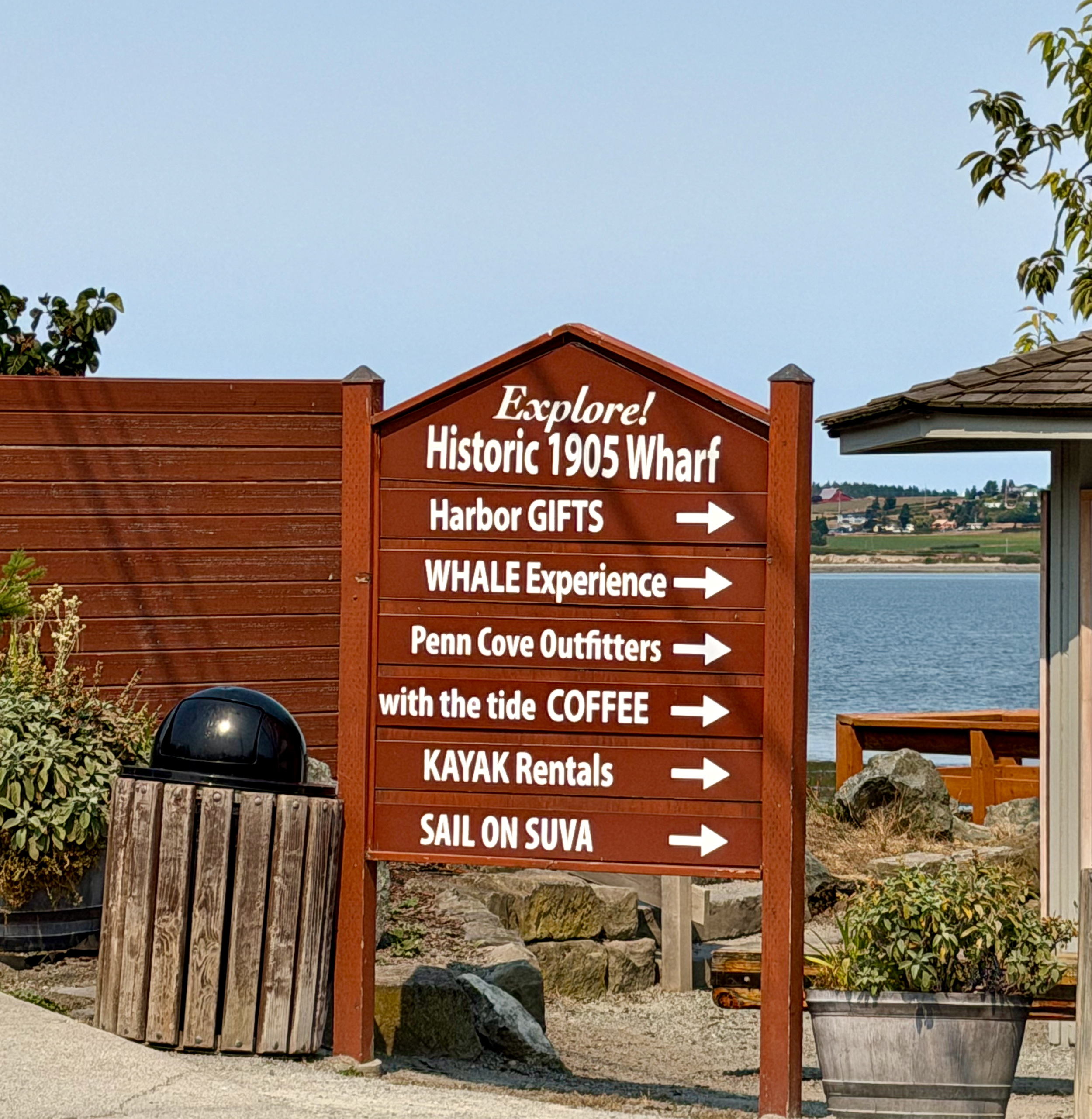 A large brown wooden sign with white text and white arrows, promoting local attractions and activities near a waterfront, including a historic wharf, harbor gifts, whale experience, kayak rentals, and sailing on the Suva.