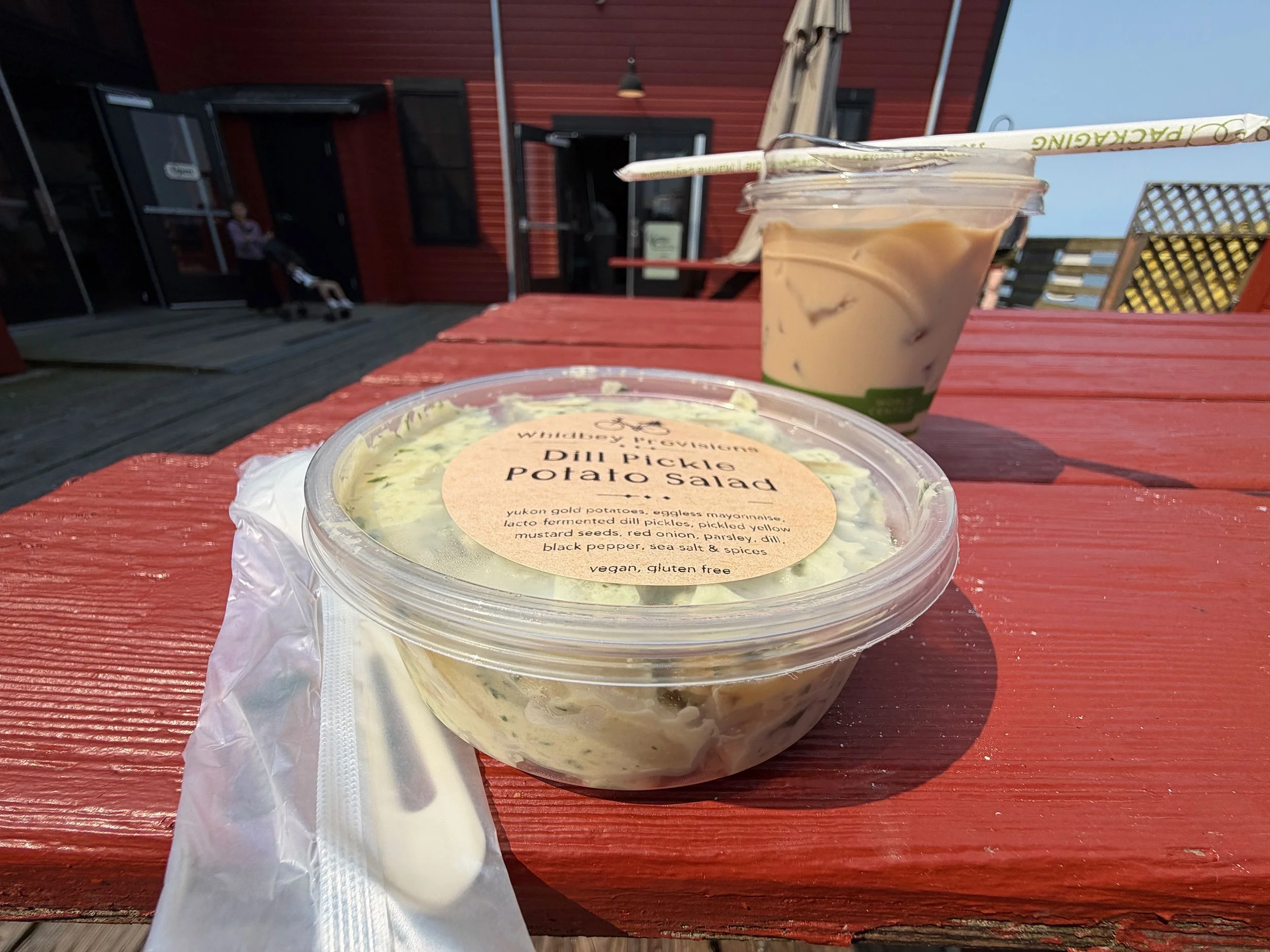 A plastic container of Yukon gold potato salad with dill pickles, eggless mayo, yellow mustard seeds, red onion, parsley, dill, black pepper, sea salt, and spices, on a red wooden table outside. A cup of iced coffee or latte with a straw is beside it