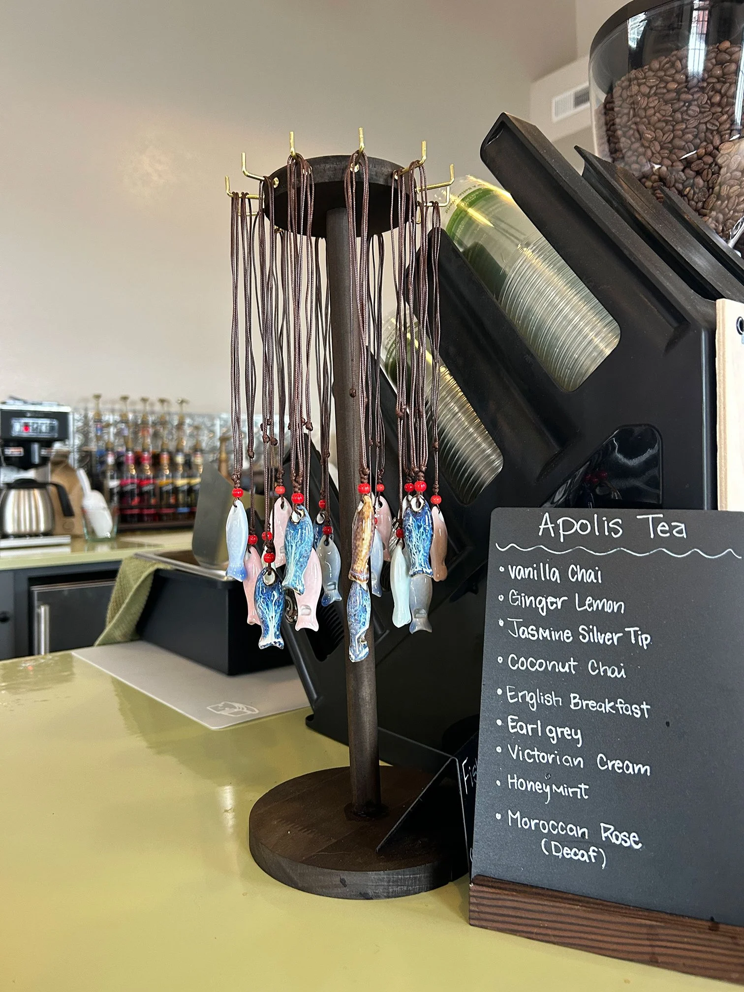 Display of fish-shaped ornaments hanging from a wooden stand with a menu on a chalkboard listing various teas, including vanilla chai, ginger lemon, jasmine silver tip, coconut chai, English breakfast, Earl Grey, Victorian cream, honey mint, Moroccan