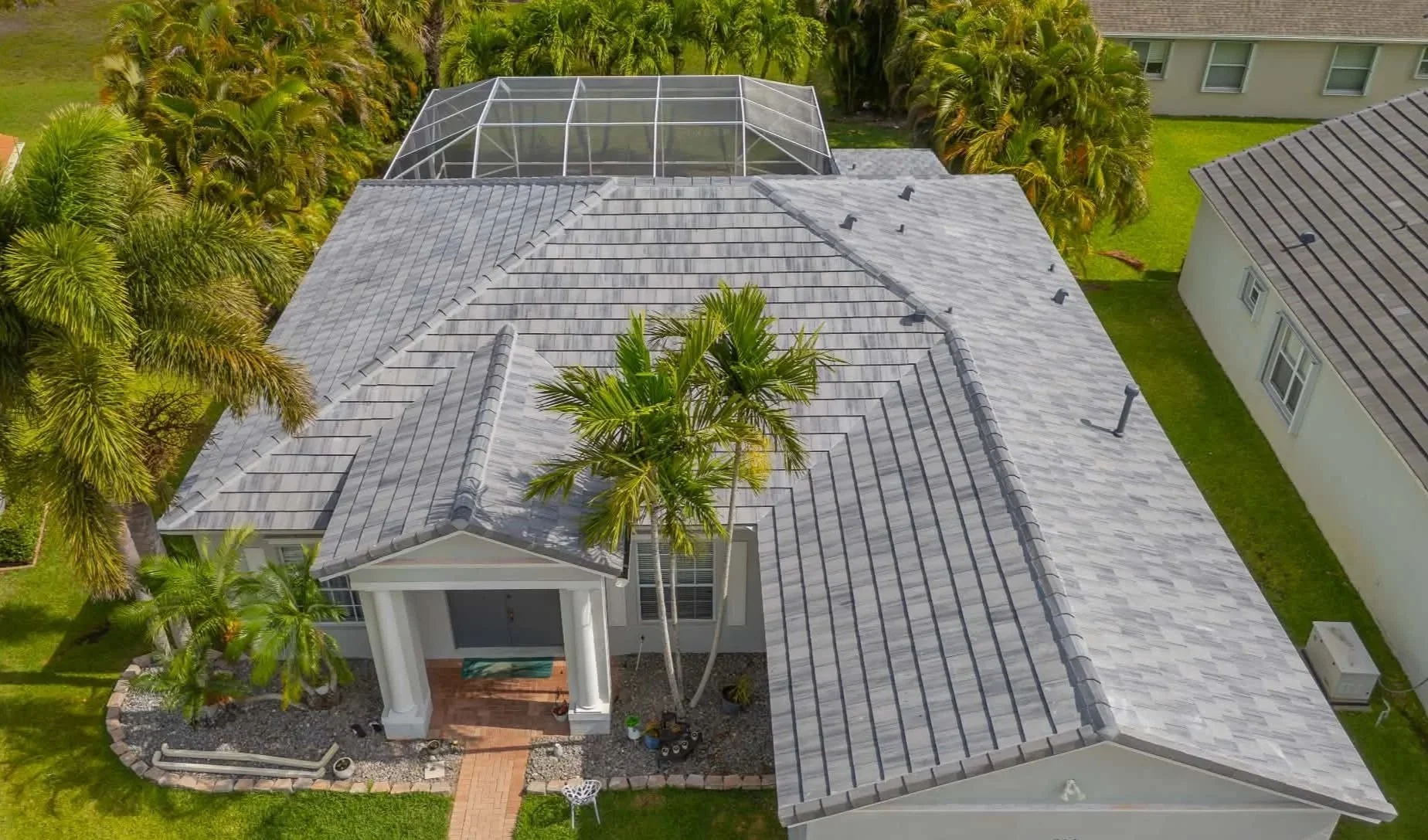 Aerial view of a house with a gray tiled roof, surrounded by palm trees, neighboring houses, and a green lawn. A screened-in pool enclosure is visible on the roof.