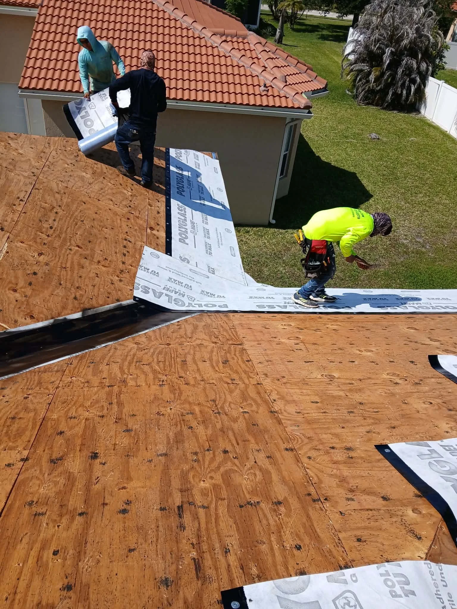 Two workers installing roof underlayment on a residential roof with plywood sheathing, outdoor yard, and trees in background.
