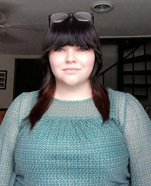 A young woman with shoulder-length dark hair with red tips, wearing sunglasses on her head, and a green patterned top, standing indoors in front of a staircase.