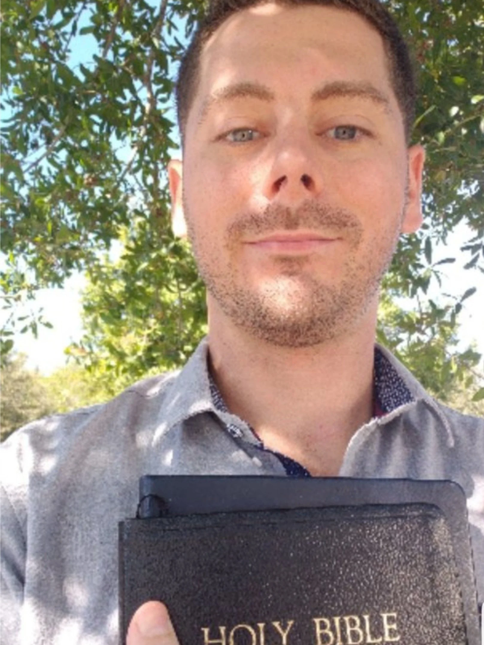 A man holding a Bible outdoors under a tree, looking at the camera.