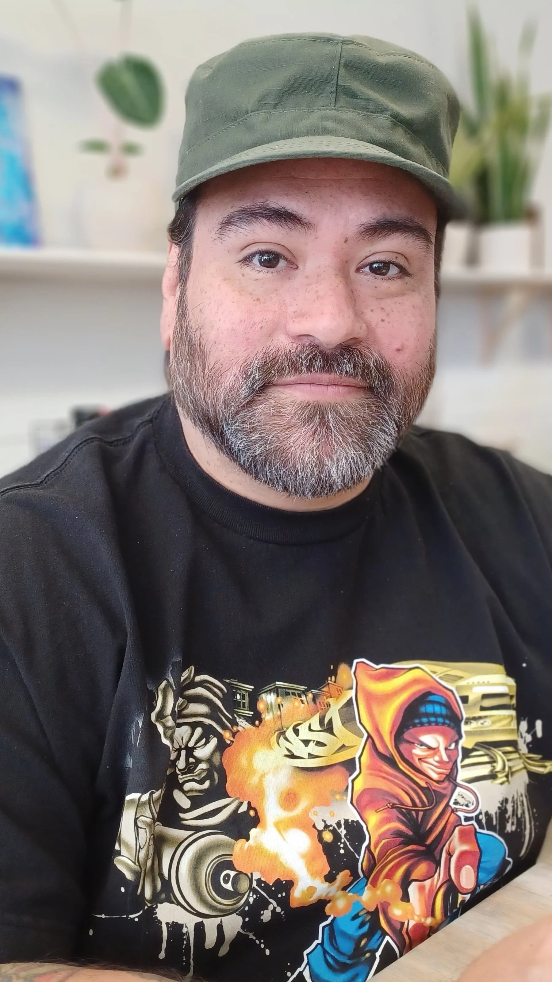 A man with a beard and mustache wearing a green cap and a black graphic T-shirt, sitting at a table in a room with white walls, plants, and a shelf in the background.