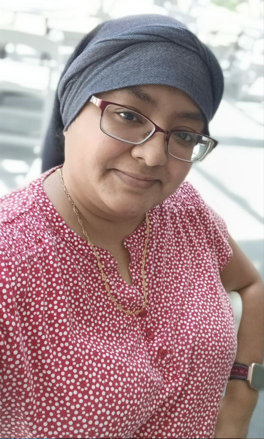 A woman with glasses, wearing a blue headscarf and a red polka dot shirt, smiling for a selfie in an indoor setting with natural light.
