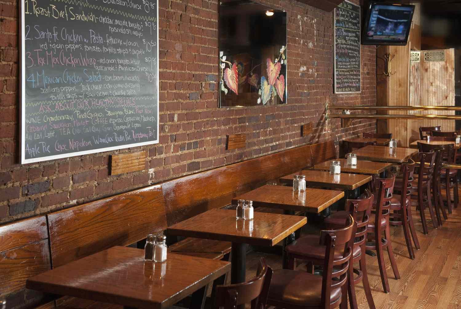 Interior of a restaurant with wooden tables, chairs, exposed brick walls, and a chalkboard menu listing food and drink options.