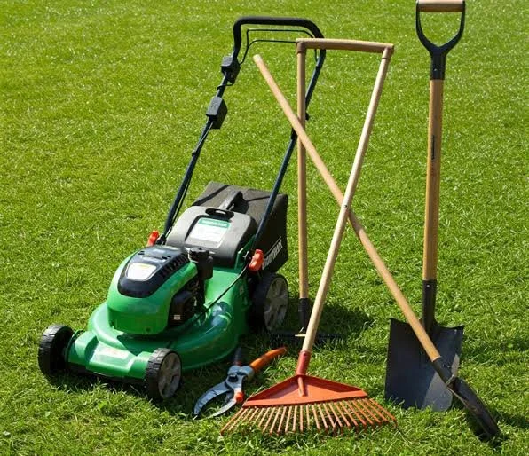 Lawn care tools on grass, including a green lawn mower, a rake, a shovel, and a post hole digger with a black handle.