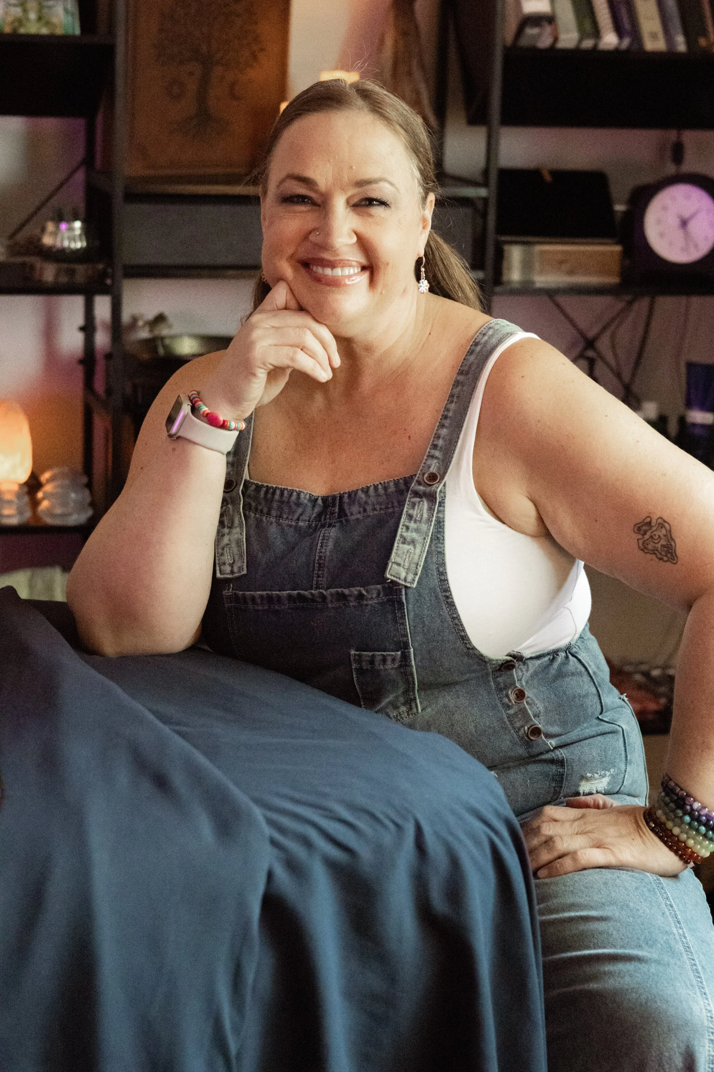A smiling woman with brown hair in a ponytail, wearing a white tank top and denim overalls, sitting indoors with a shelf of books and a clock in the background.