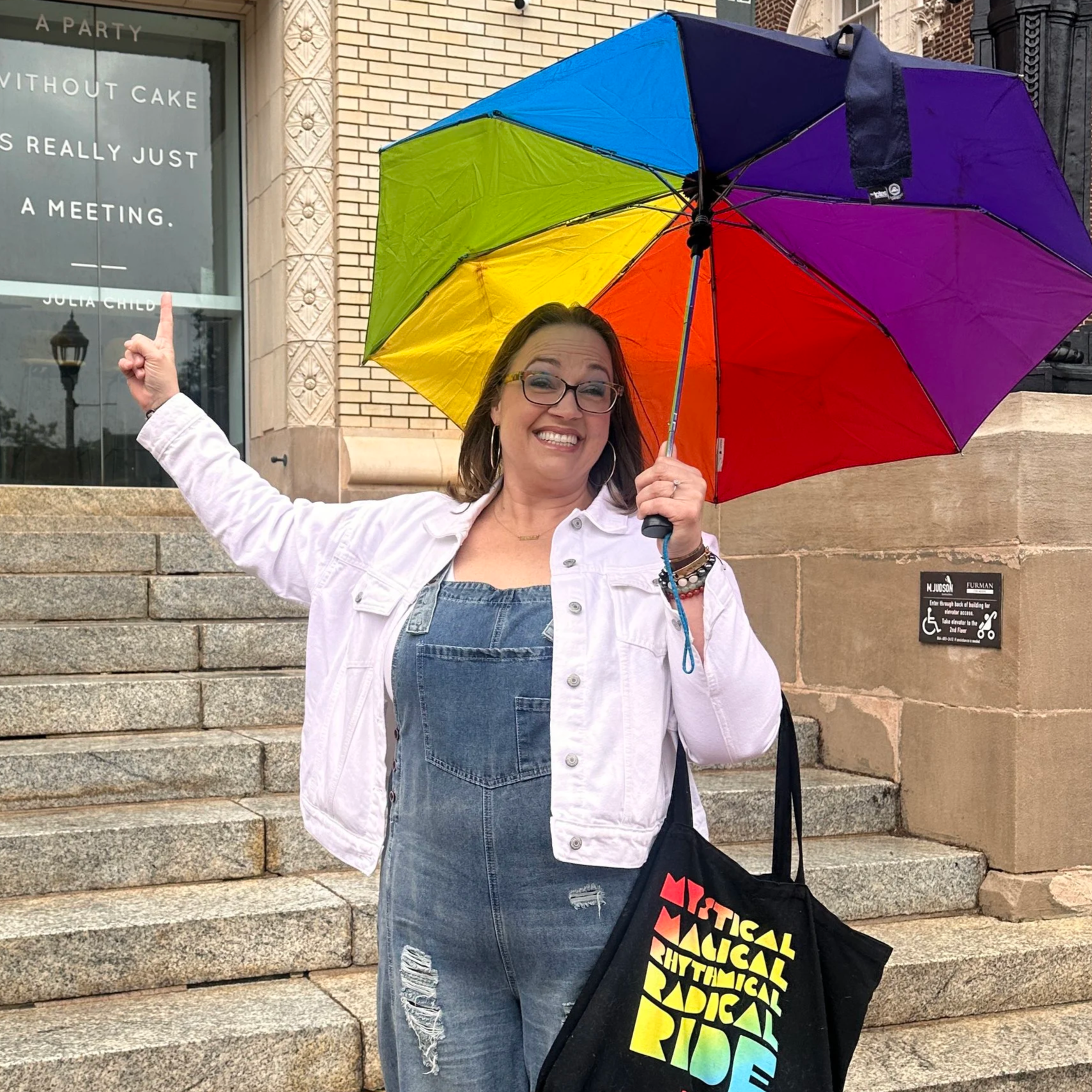 A woman standing outside on a set of stone stairs holding a rainbow-colored umbrella. She is smiling, pointing upward with her right hand, and wearing glasses, hoop earrings, a white jacket, a denim dress, and several bracelets. In the background, there is a brick building, a window with text, and a black bag with rainbow text that says 'Material Racism Systemic.'