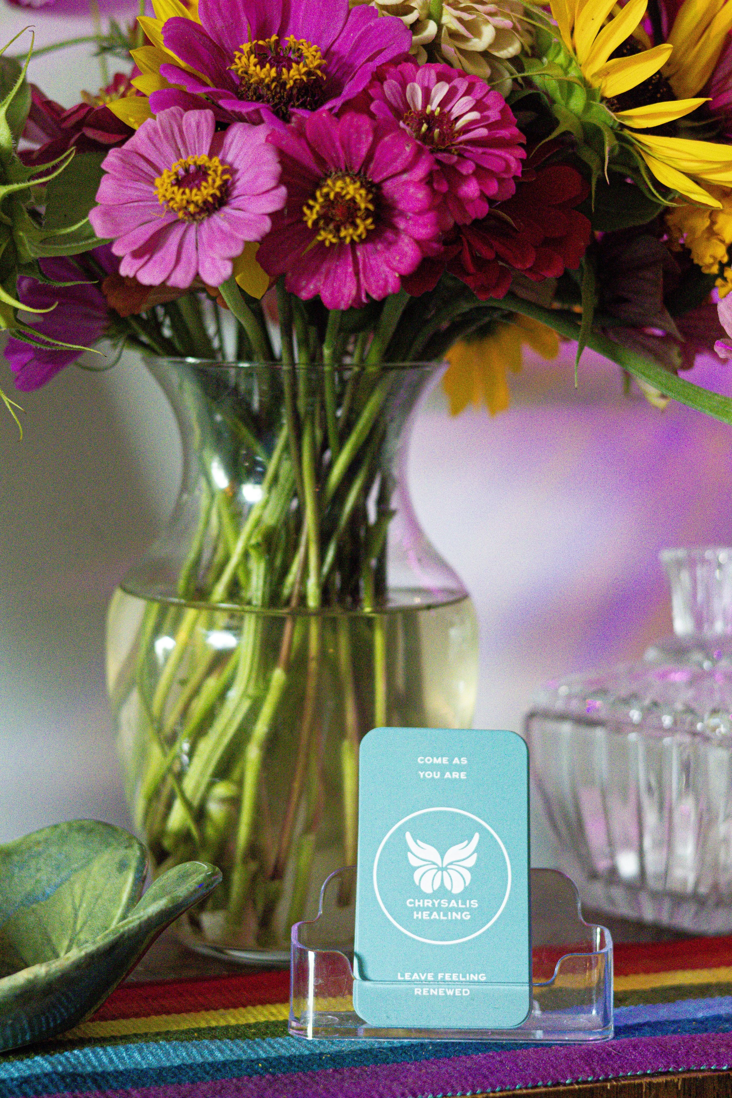 A colorful bouquet of flowers in a glass vase on a wooden surface, with a symbol of a butterfly and the words 'Chrysalis Healing' on a card in front, and a rainbow-striped fabric underneath.