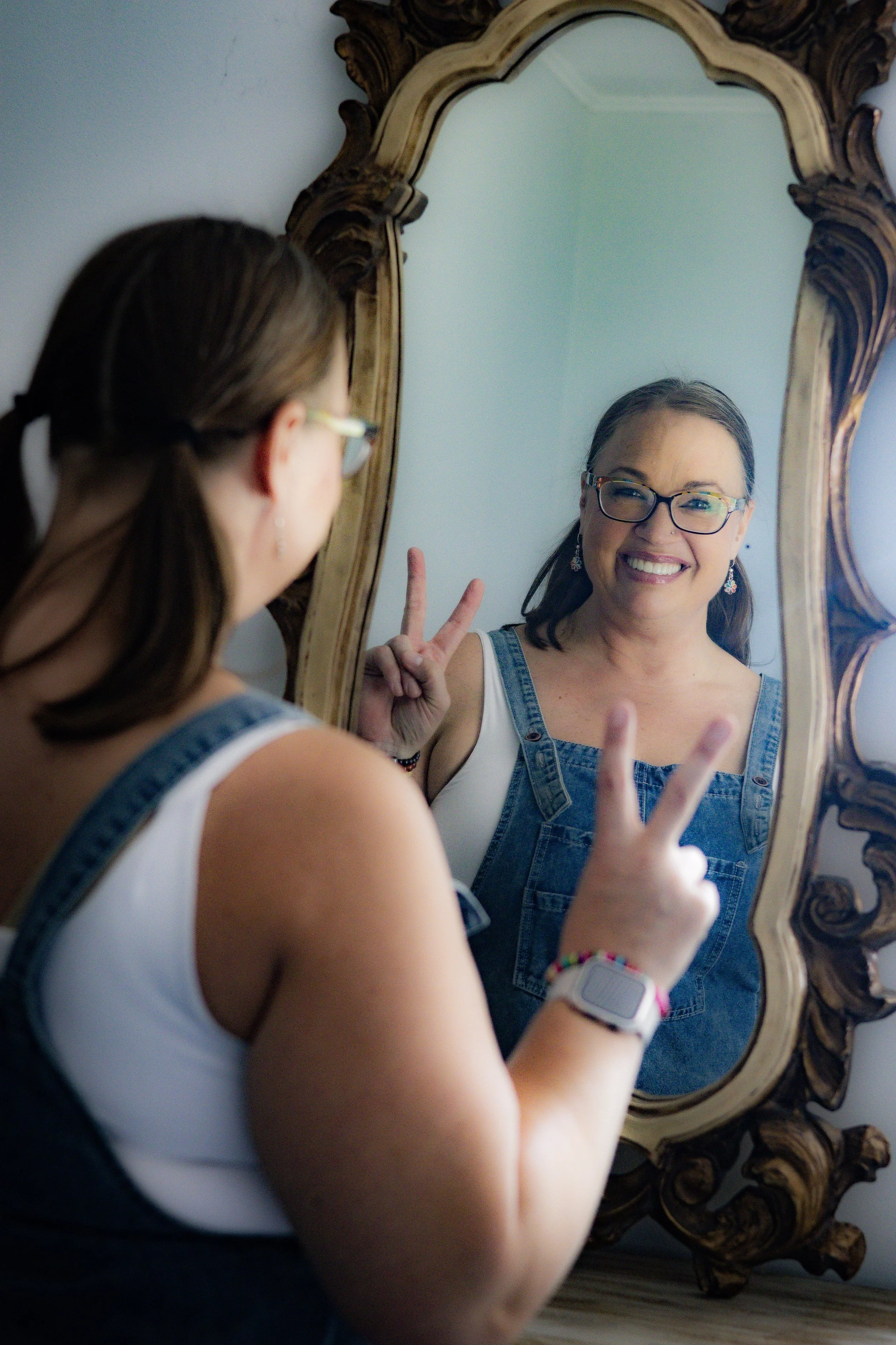 Smiling woman with glasses and earrings taking a selfie in a mirror, making a peace sign with her fingers, standing in front of an ornate wooden mirror frame.