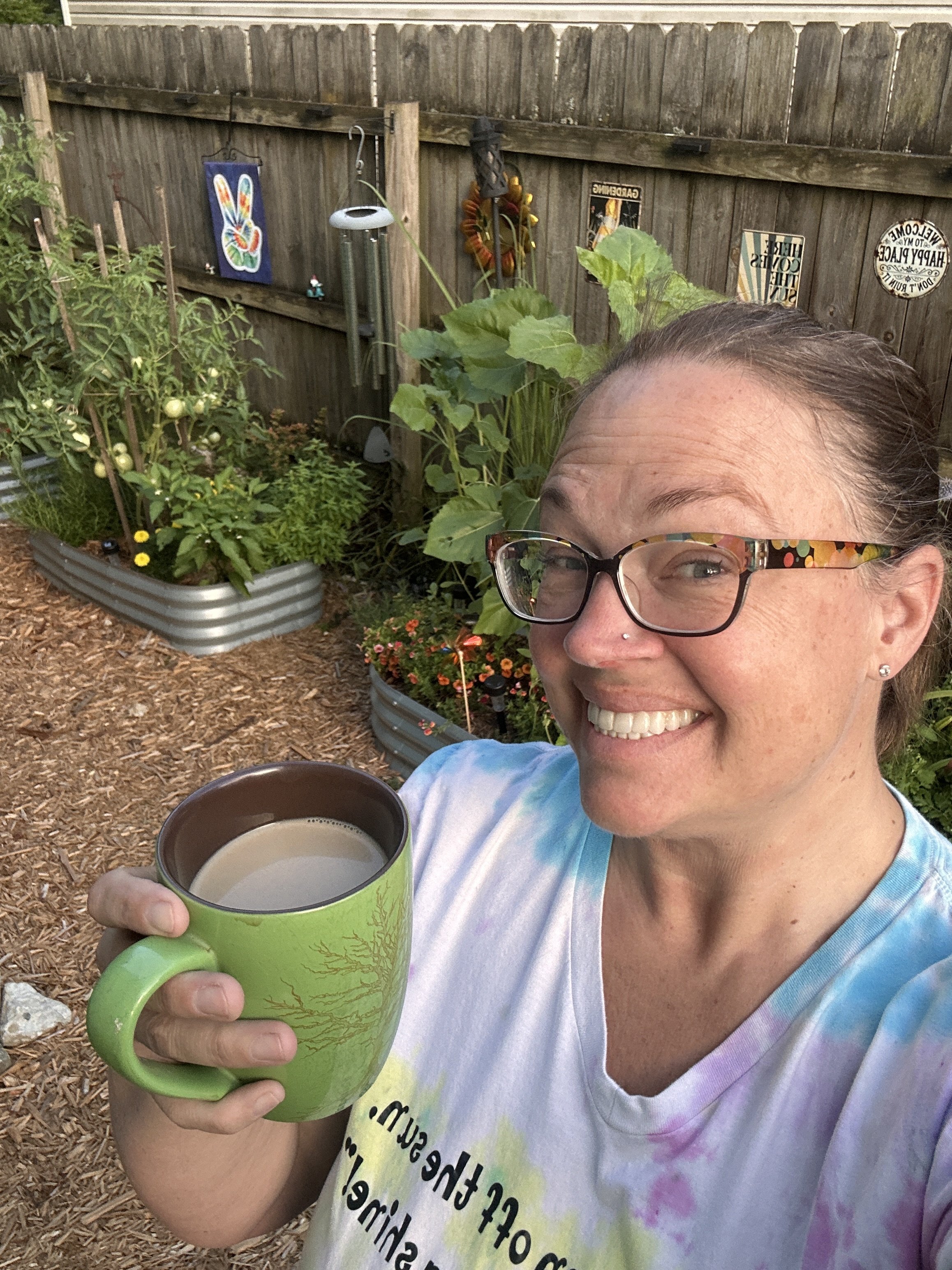 A woman smiling outdoors, holding a green mug with a tree design, wearing glasses and a tie-dye shirt, with a garden background of plants, flowers, and decorative signs on a wooden fence.