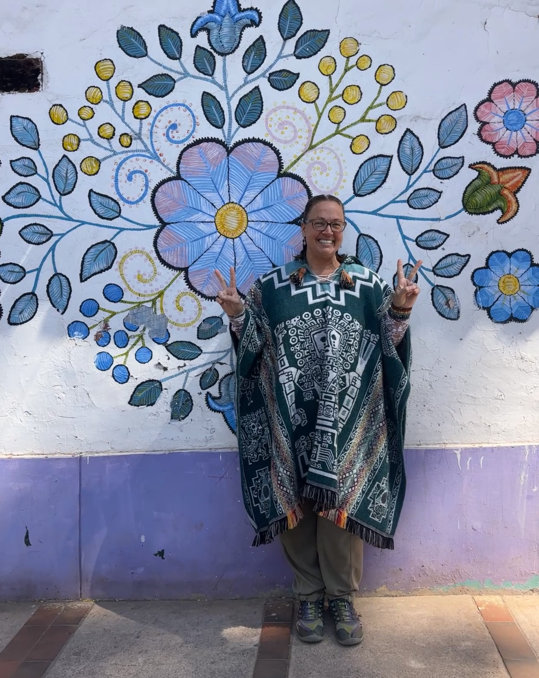 Woman standing in front of a colorful mural with flowers and leaves, smiling and making peace signs with both hands.