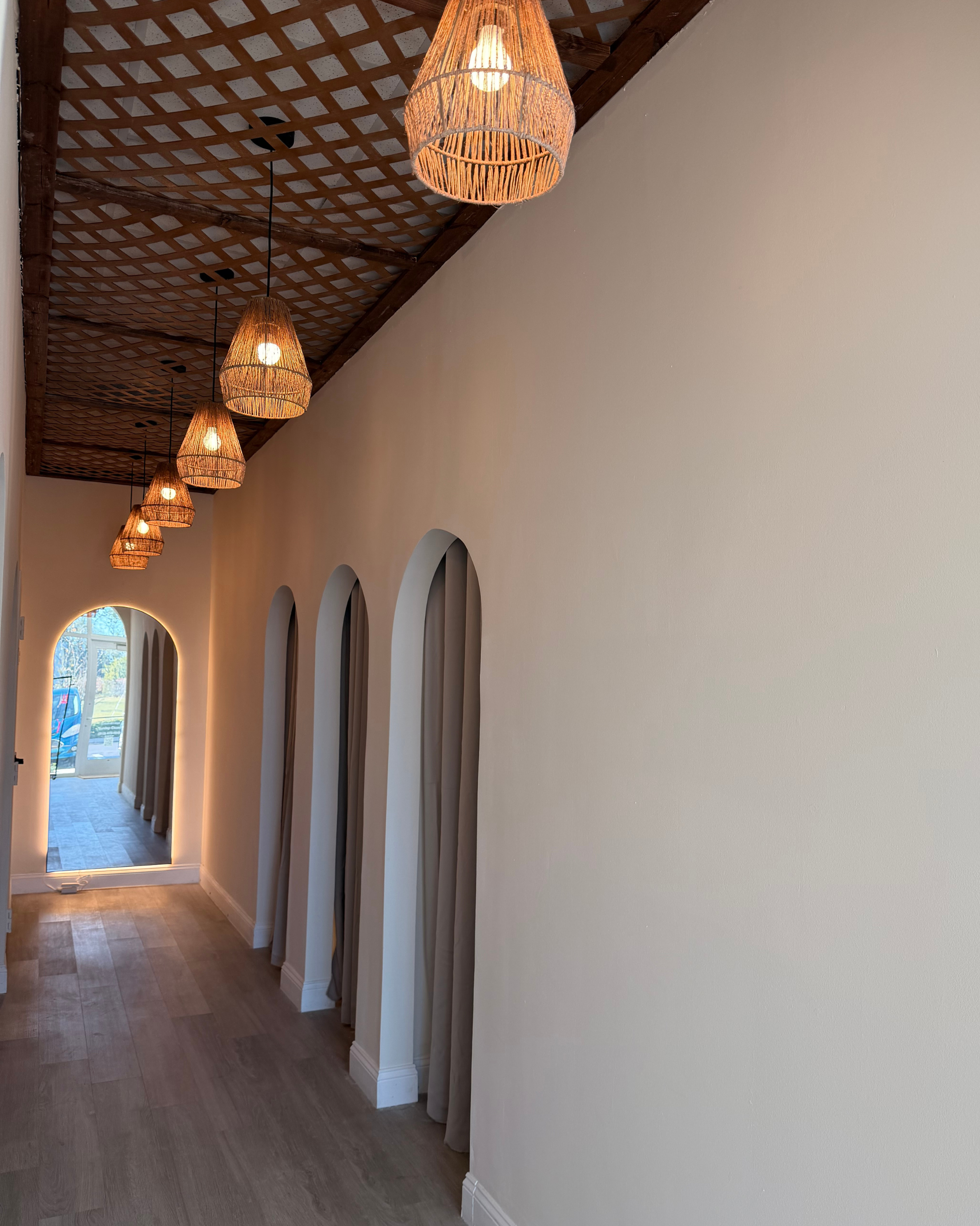 Interior hallway with a wooden lattice ceiling, hanging wicker pendant lights, arched windows along the left wall, and a door at the end with a view outside.