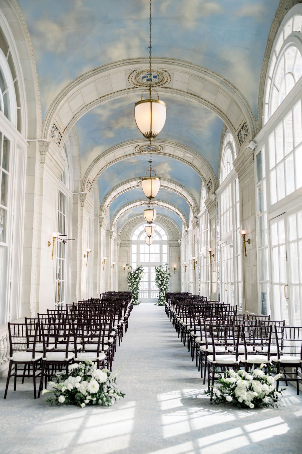 Elegant indoor wedding ceremony setup in a bright, glass-enclosed conservatory with white floral arrangements at the end of the aisle.