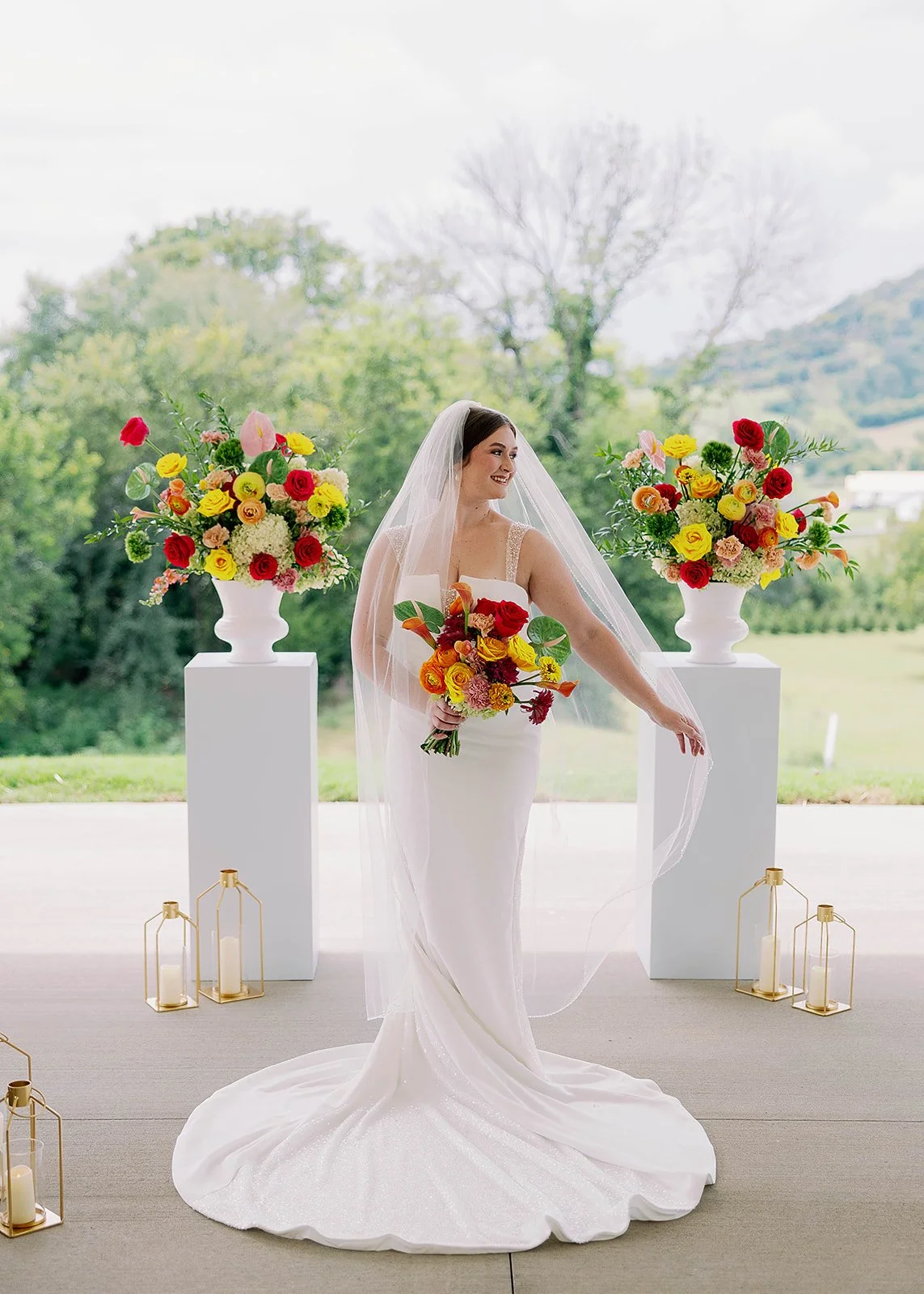 Bride in a white gown and veil holding a bouquet of colorful flowers, standing between two large floral arrangements on white pedestals, with candles in gold lanterns on the floor, against an outdoor backdrop of trees and hills.