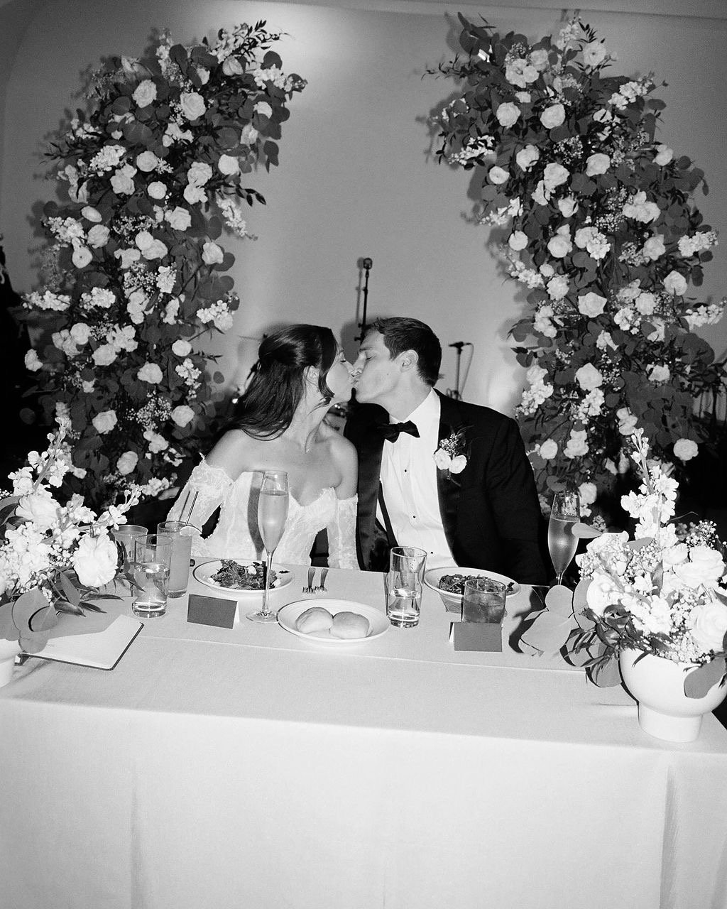 A black and white photo of a bride and groom sharing a kiss at their wedding reception. They are seated at a table decorated with floral arrangements and surrounded by large floral arches.