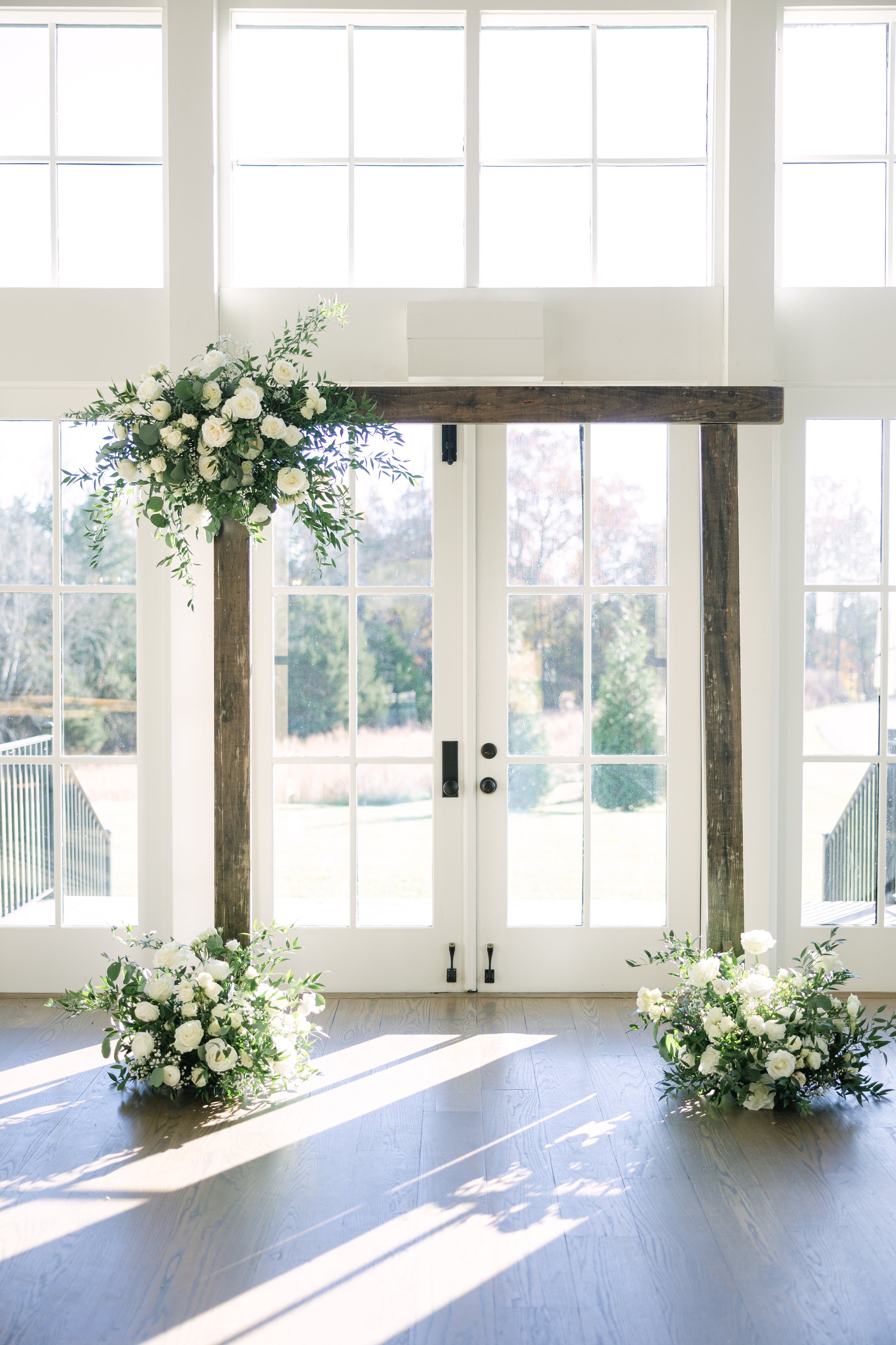 Elegant white floral wedding arch with greenery in front of large windows with sunlight streaming in.