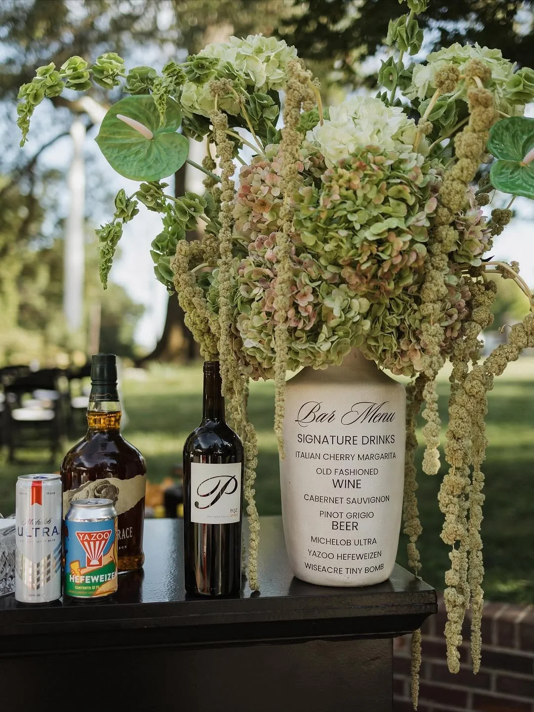 A floral arrangement with green hydrangeas and greenery, along with bottles of wine, beer, and cans on a black table, outdoors in a park-like setting.
