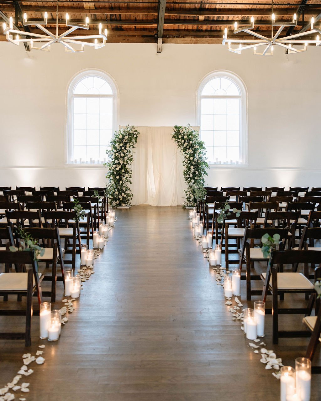 Wedding aisle decorated with white candles and rose petals leading to a floral arch at the front of a bright, elegant ceremony space with large arched windows.