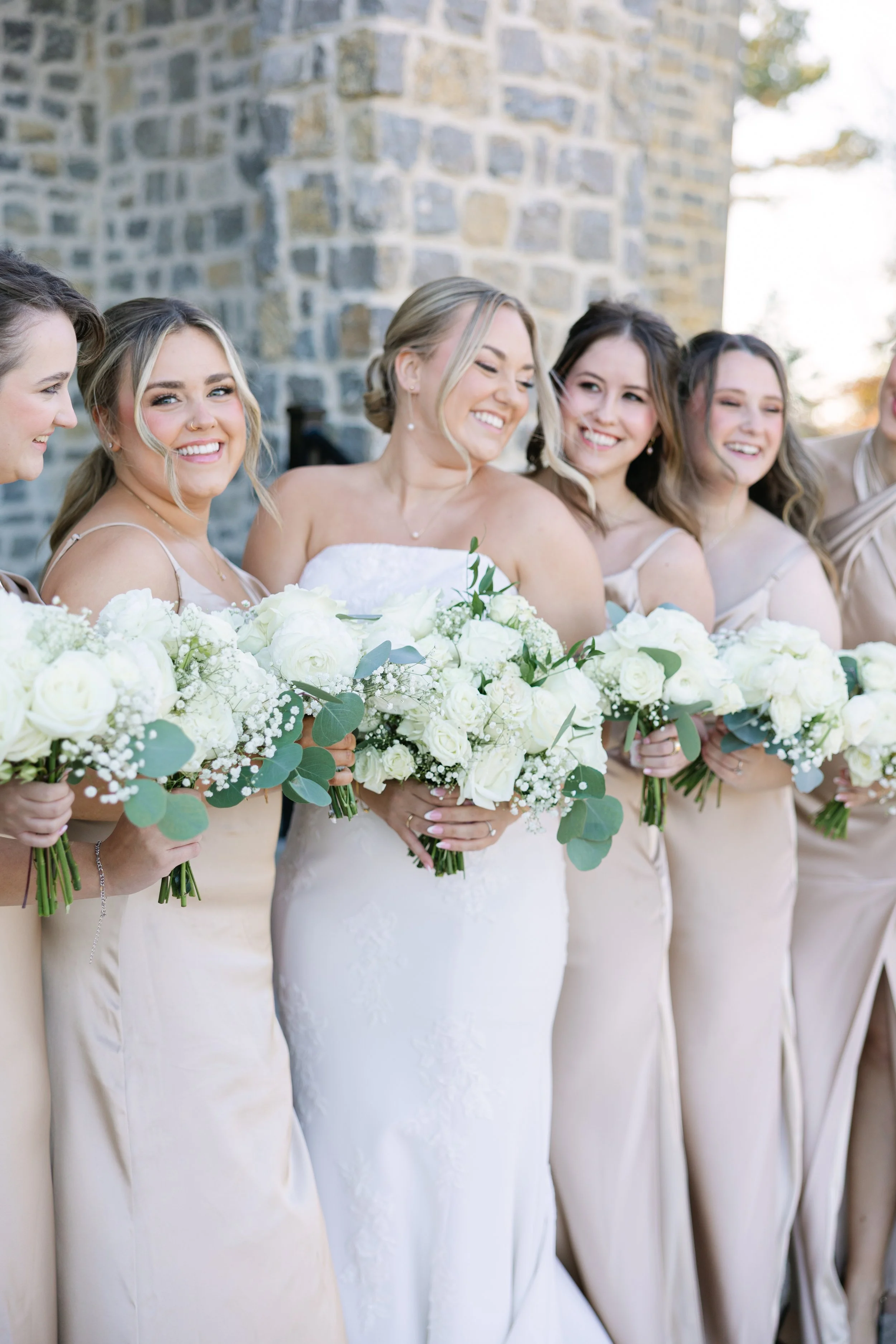 A bride and five bridesmaids standing together outdoors, smiling, holding bouquets of white roses and greenery, with a stone wall in the background.