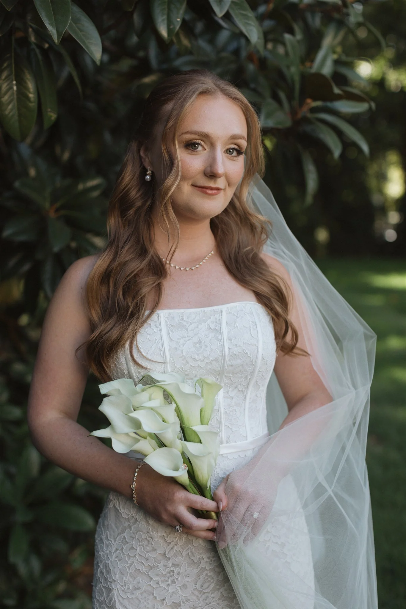 A bride with long wavy brown hair is holding a bouquet of white calla lilies. She is wearing a strapless white lace wedding dress, pearl earrings, a pearl necklace, and a bracelet. She is outdoors with green foliage in the background, smiling softly.