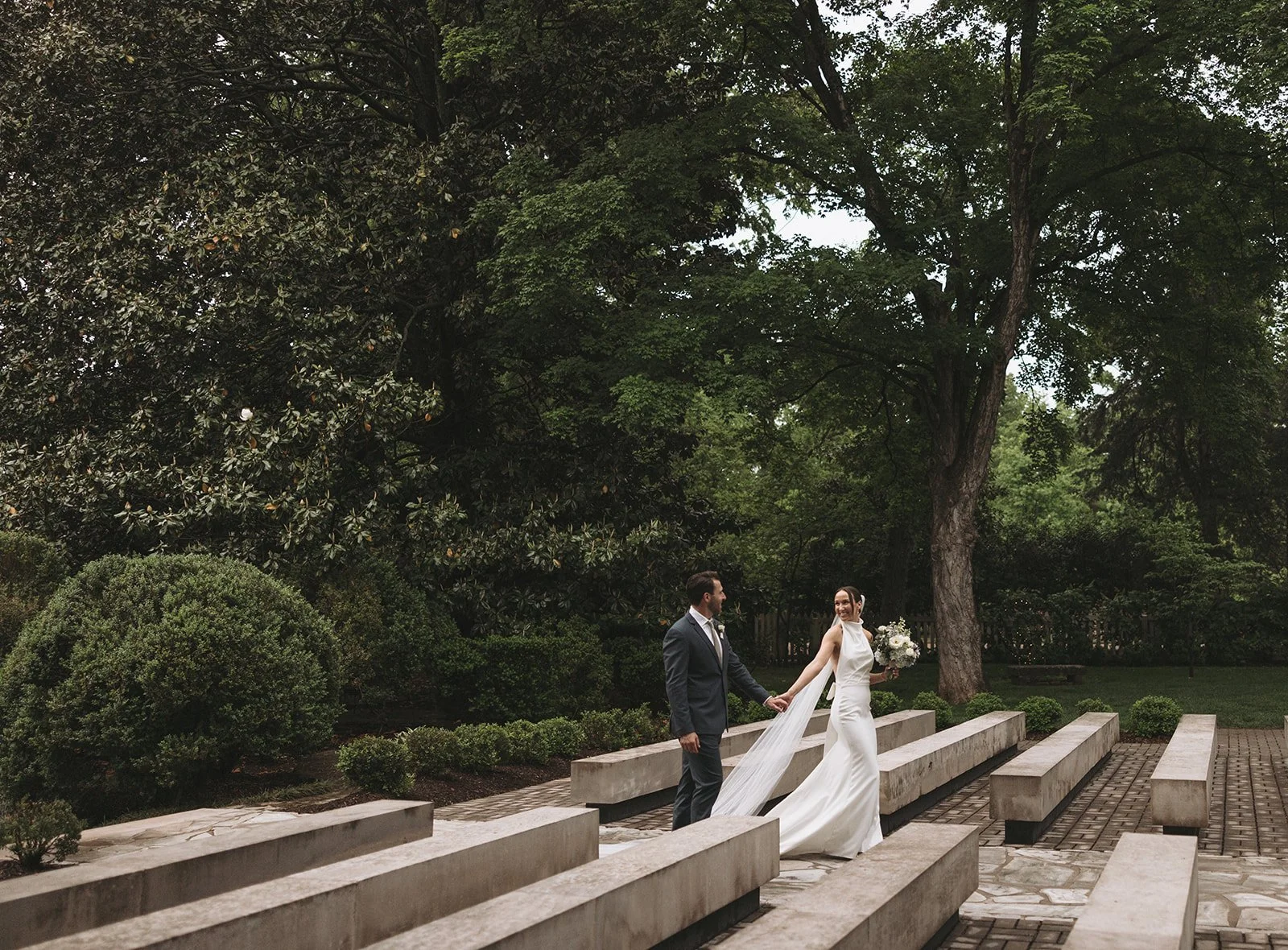 A bride and groom holding hands on a stone staircase outdoors surrounded by lush green trees and bushes, during daytime.