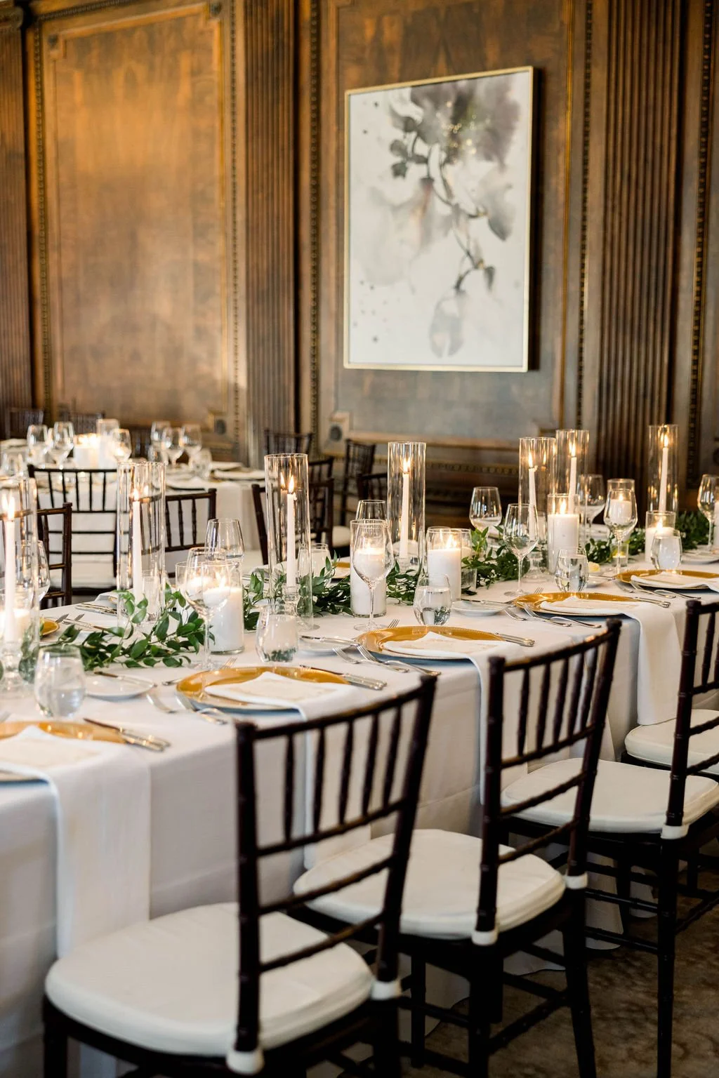 Elegant dining table set for a formal event with white tablecloths, gold-rimmed plates, silverware, glassware, and green foliage garland. Tall candles and lanterns as centerpieces are on the table. Wooden chairs surround the table. The background fea