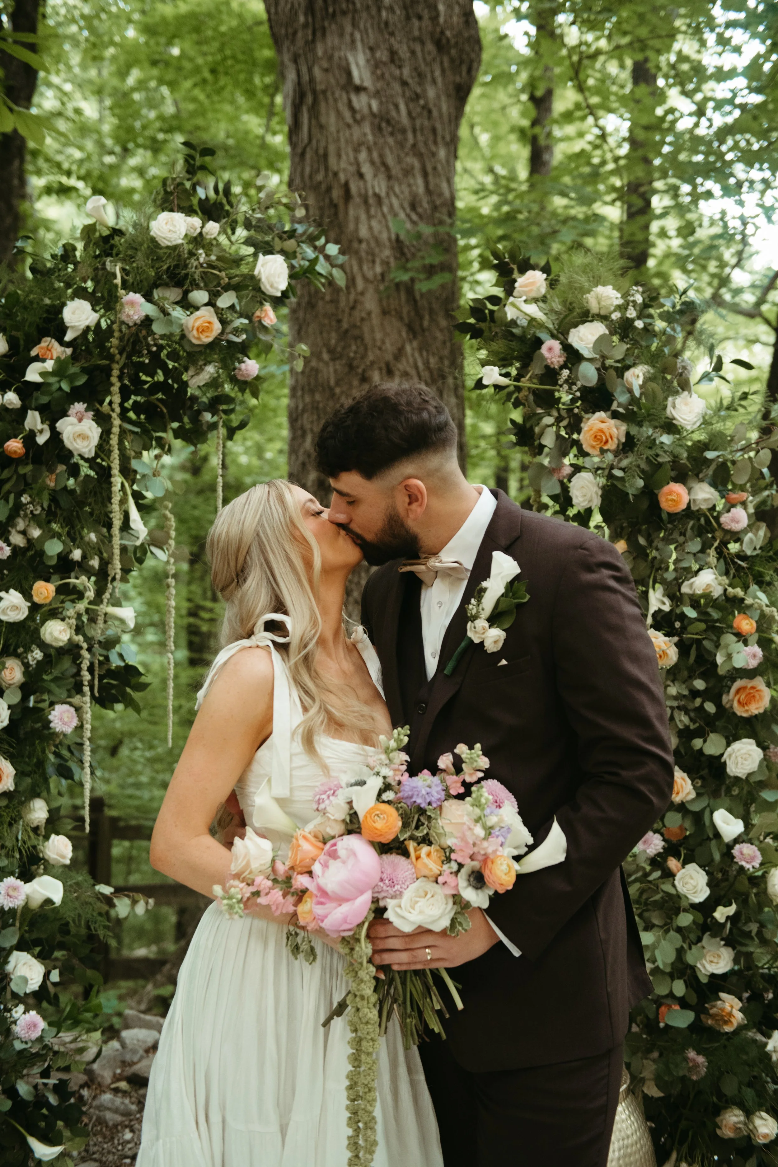 A bride and groom kissing during their outdoor wedding ceremony in a forest decorated with flowers and greenery.