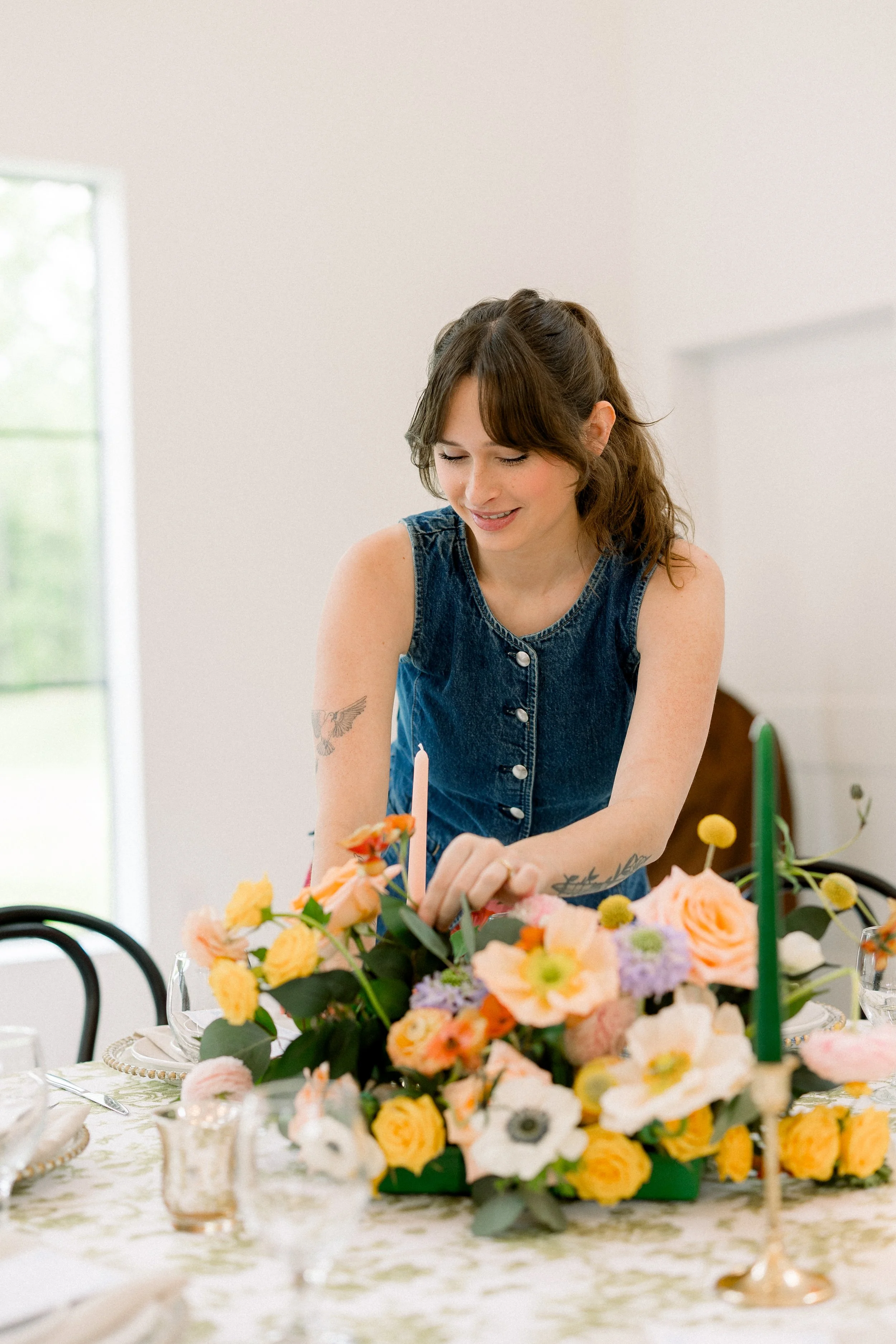 A woman in a denim sleeveless top arranging a floral centerpiece at a table set for a celebration or gathering.