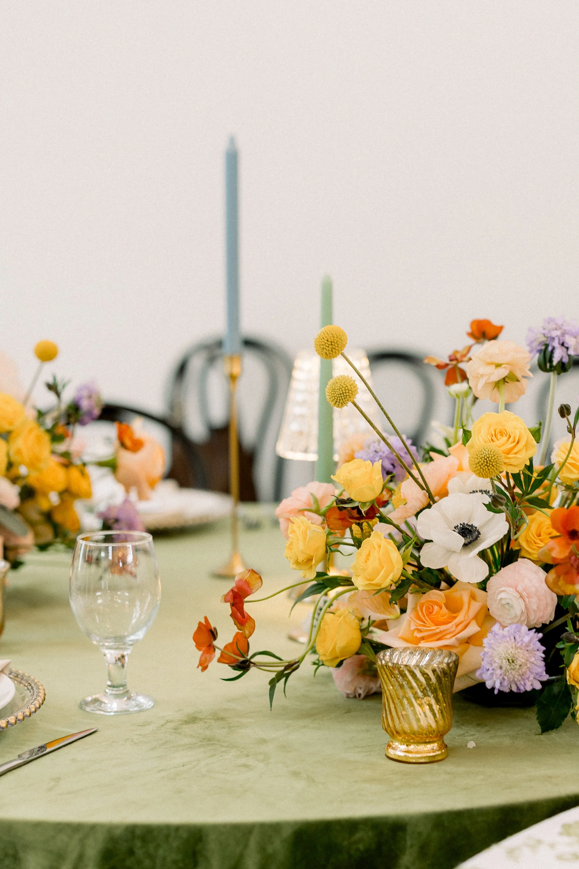 A table with a large floral arrangement in a gold vase, two tall candles, and empty wine glasses, set for a formal occasion.