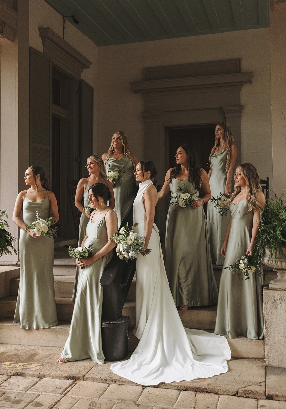 A group of nine women dressed in matching satin gowns, holding bouquets, standing on the steps of a building during a wedding or special event.