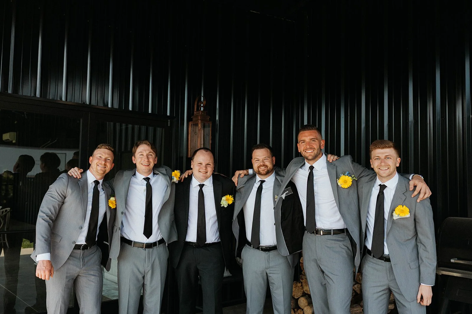 Group of seven men in suits with yellow flower boutonnieres, smiling with arms around each other, standing against a black wall.