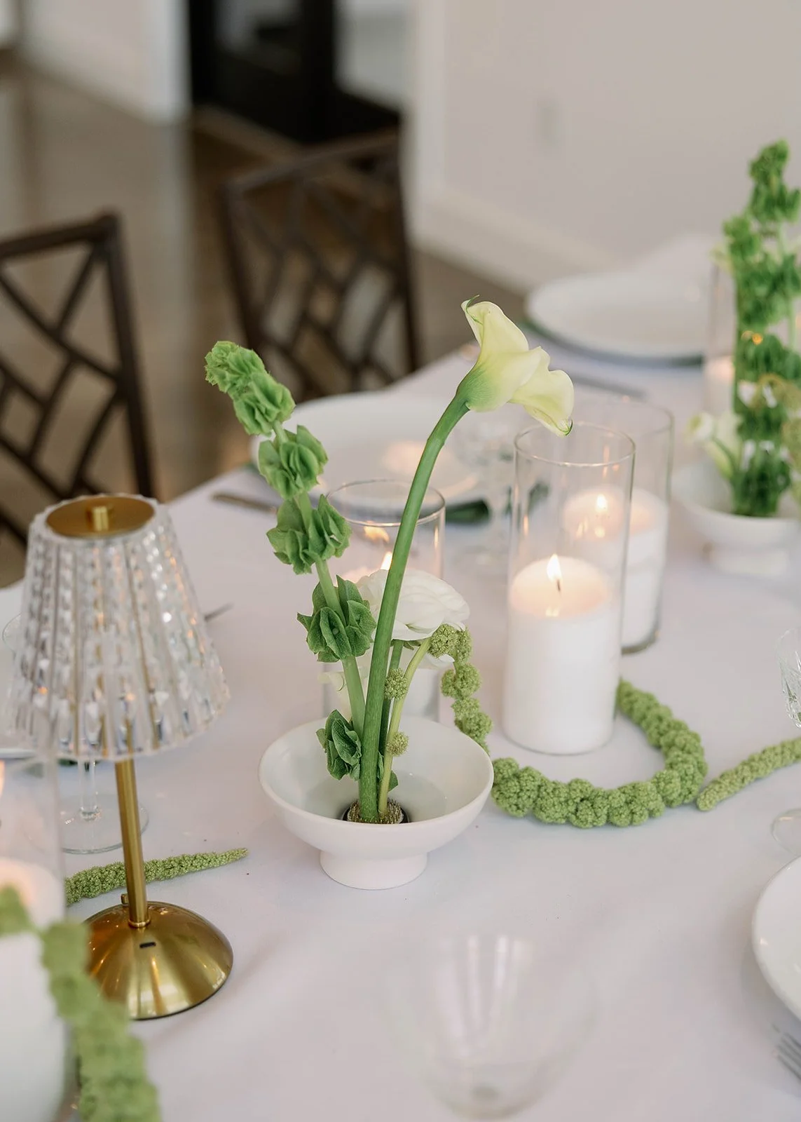 Elegant table centerpiece with white and green flowers in a small white vase, surrounded by candles in tall glass holders, a gold table lamp, and green decorative garlands on a white tablecloth.