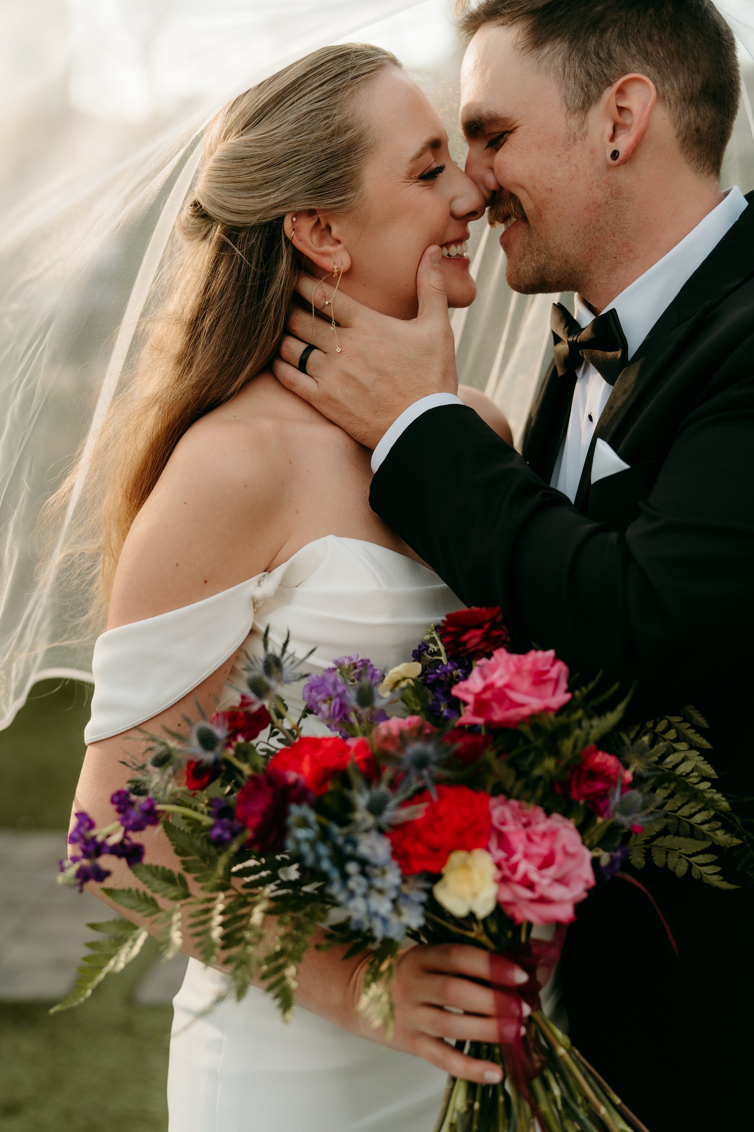 A husband and wife posing together with a jewel toned bouquet. 
