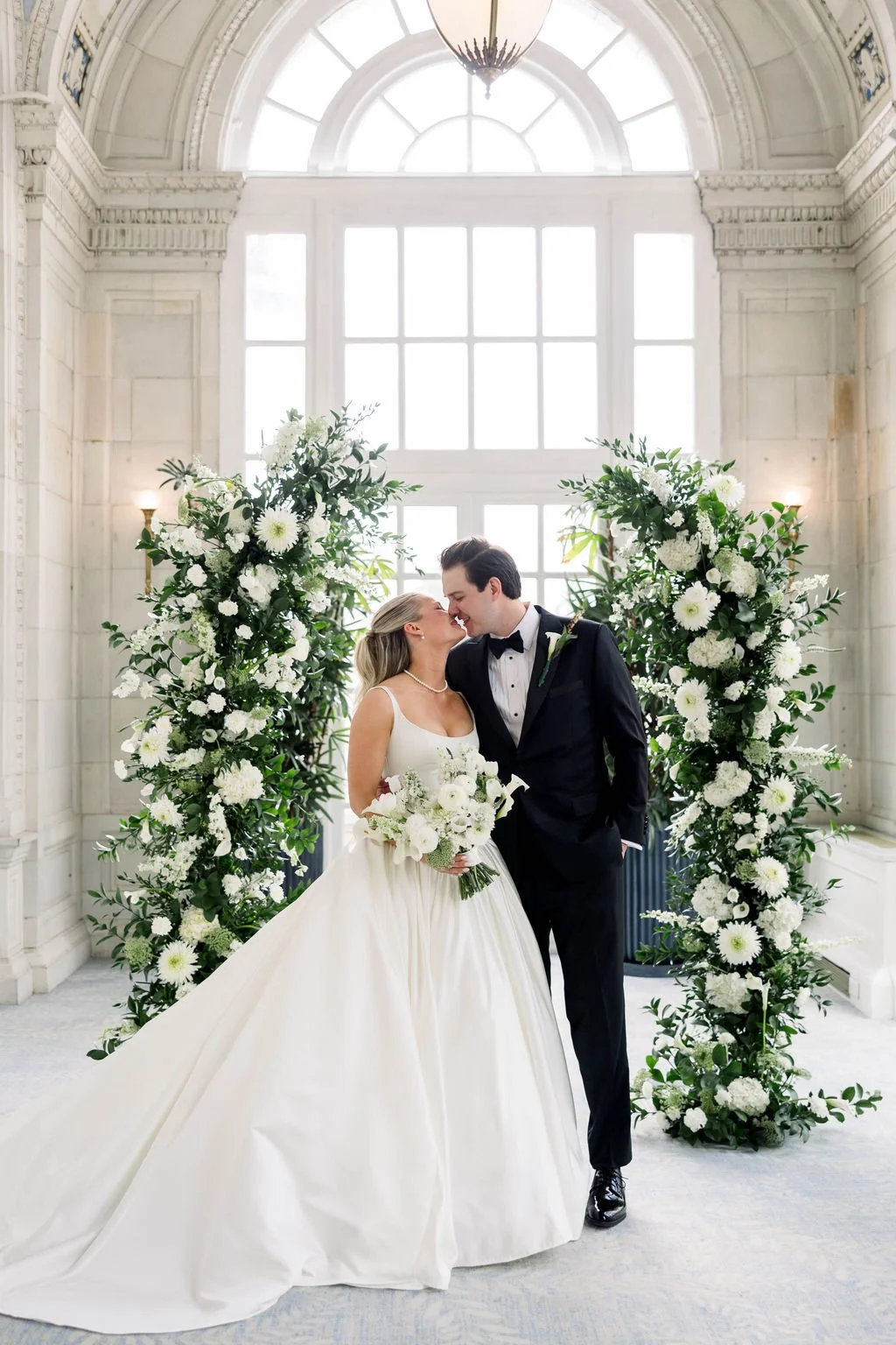 A bride and groom standing face to face and touching noses, surrounded by a large floral arch inside a bright, elegant building with high windows and chandeliers.