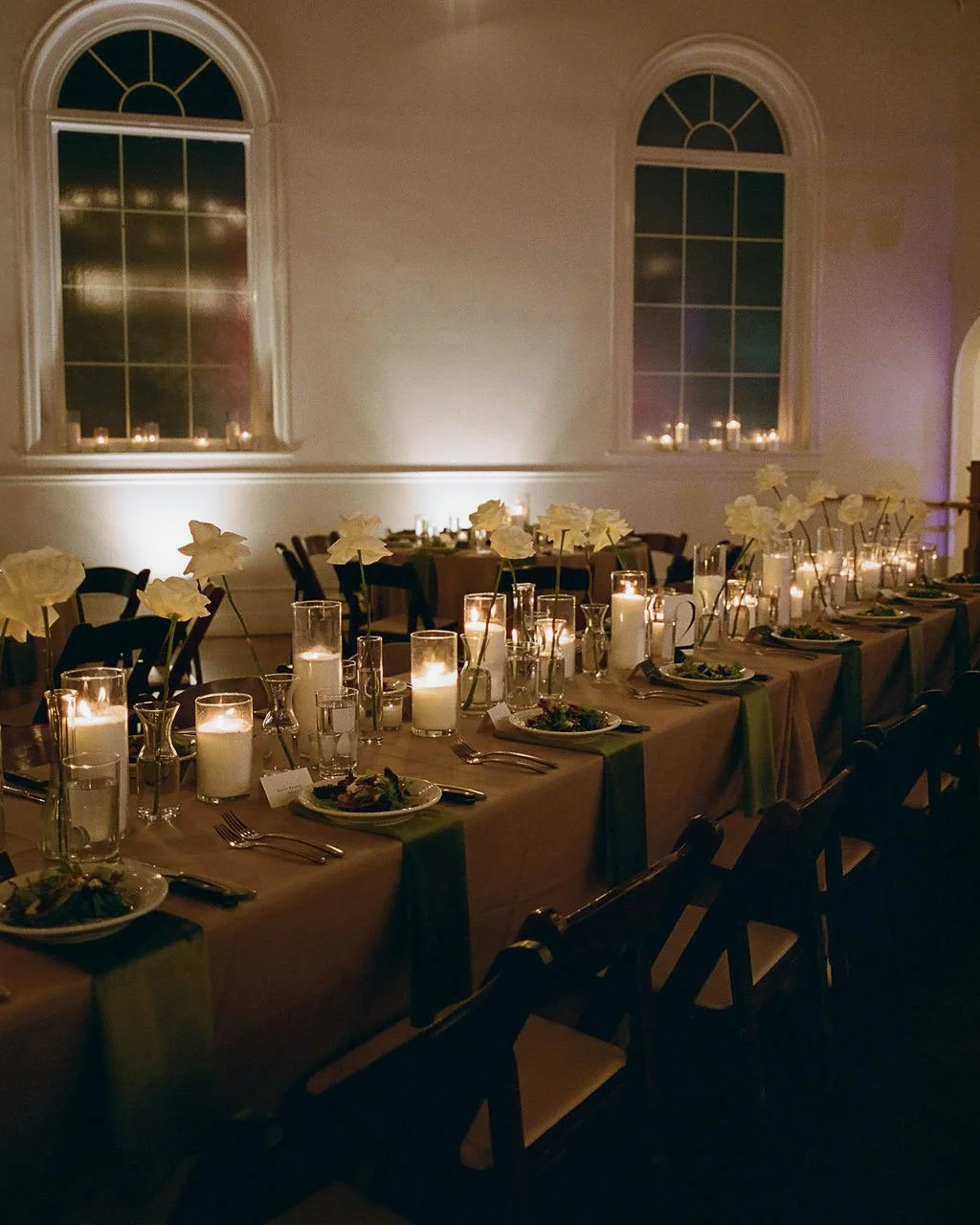 Elegant dining table decorated with white flowers, candles, and place settings in a dimly lit room with two large arched windows.