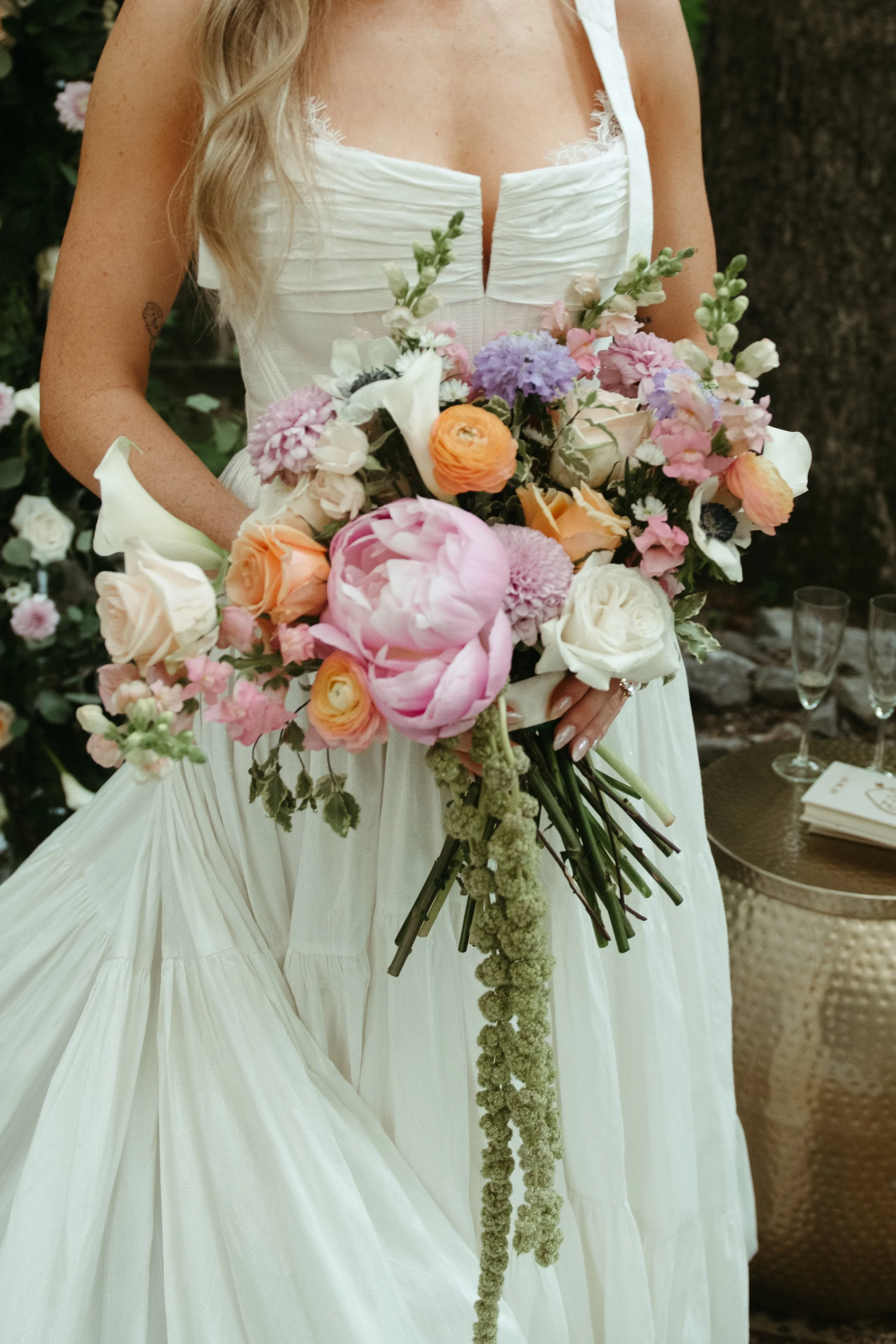 A woman in a white dress holding a large bouquet of colorful flowers, standing outdoors near a tree.