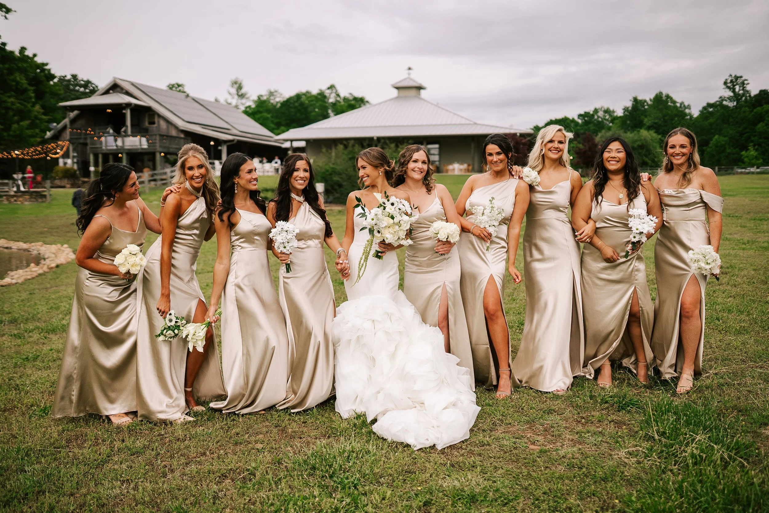 Group of nine women in matching satin dresses smiling and holding bouquets, standing outdoors on a grassy field at a wedding or celebration, with a barn and a pavilion in the background under cloudy sky.