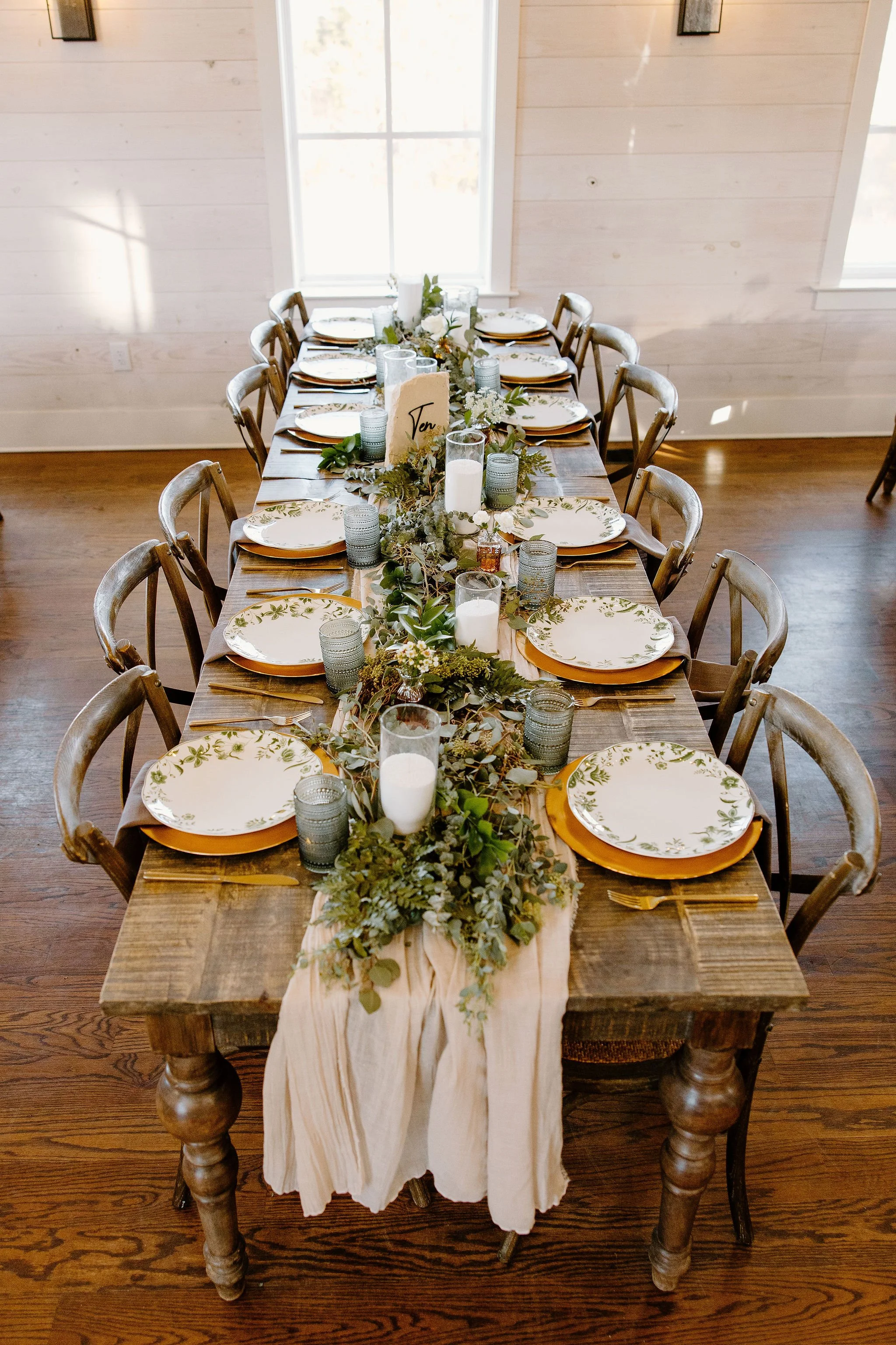 A rectangular wooden dining table set for ten people with floral-patterned plates, gold-rimmed chargers, and gold utensils. The table is decorated with a greenery and eucalyptus garland, white candles in glass holders, and champagne-colored napkins. A table number sign reading 'Ten' is placed among the candles. The table is surrounded by wooden chairs, and the room has whitewashed wooden walls and large windows with sunlight streaming in.