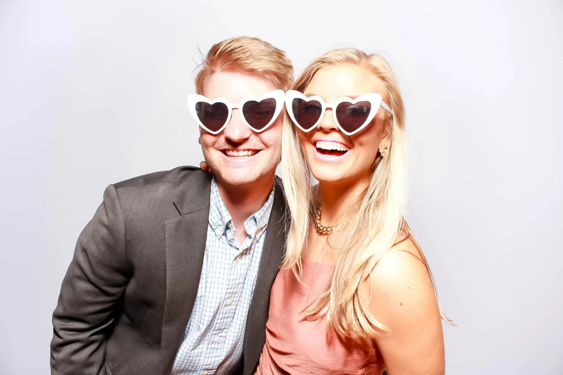 A smiling man and woman wearing large heart-shaped sunglasses, dressed casually, standing close together against a plain light gray background.