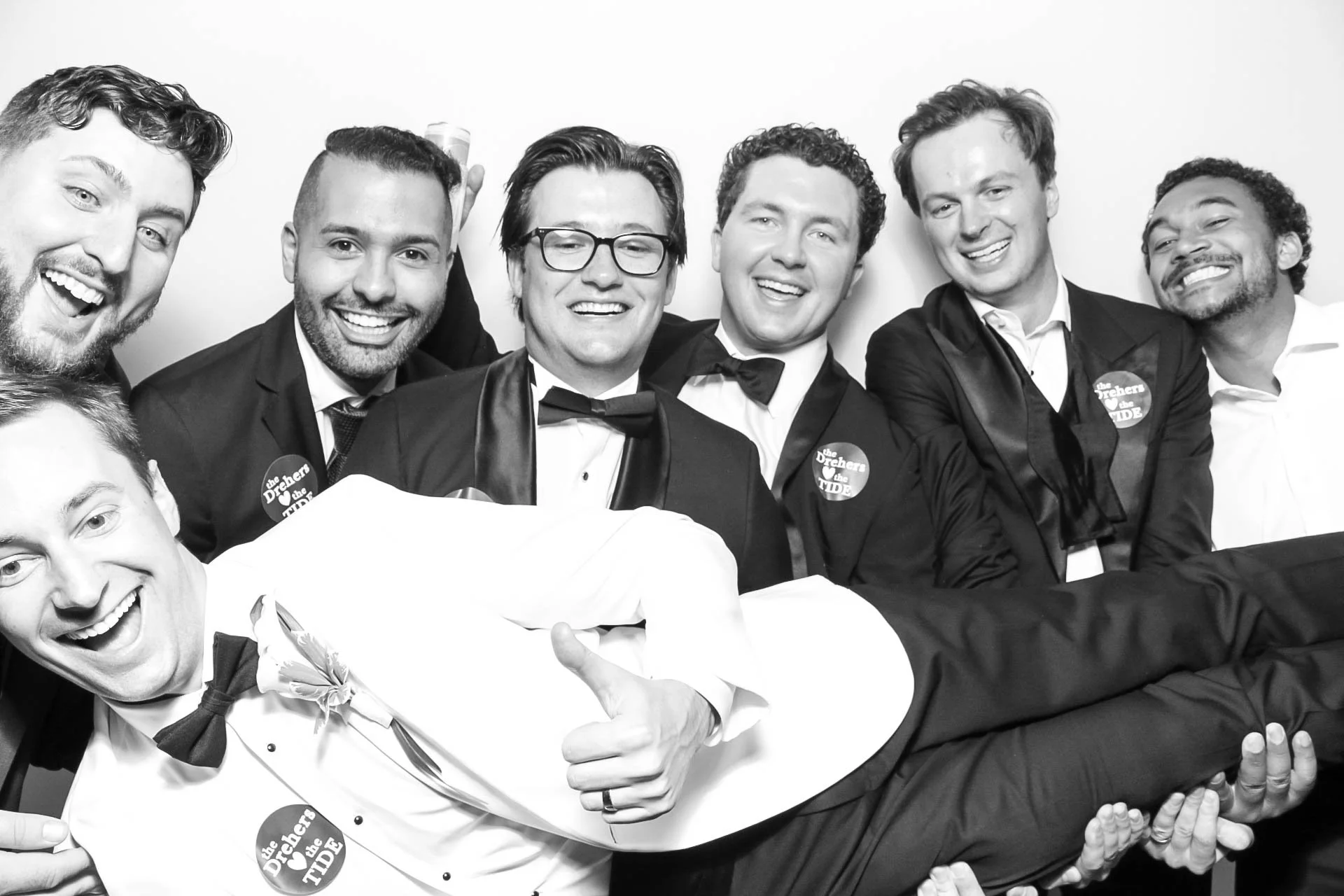 Group of six men wearing tuxedos and bow ties, smiling and celebrating at a formal event, some have badges that the couple loves the Alabama Crimson tide.