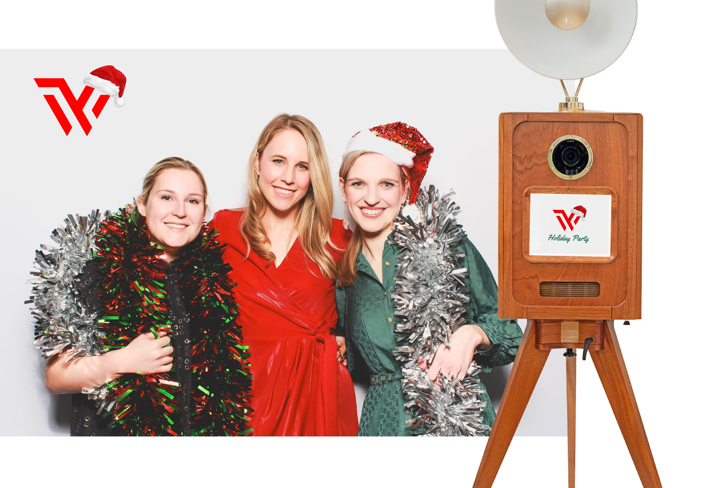 Three women in Christmas hats and festive clothing posing with silver tinsel and a wood speaker with a small screen that reads "Holiday Party" and a logo with a Santa hat.